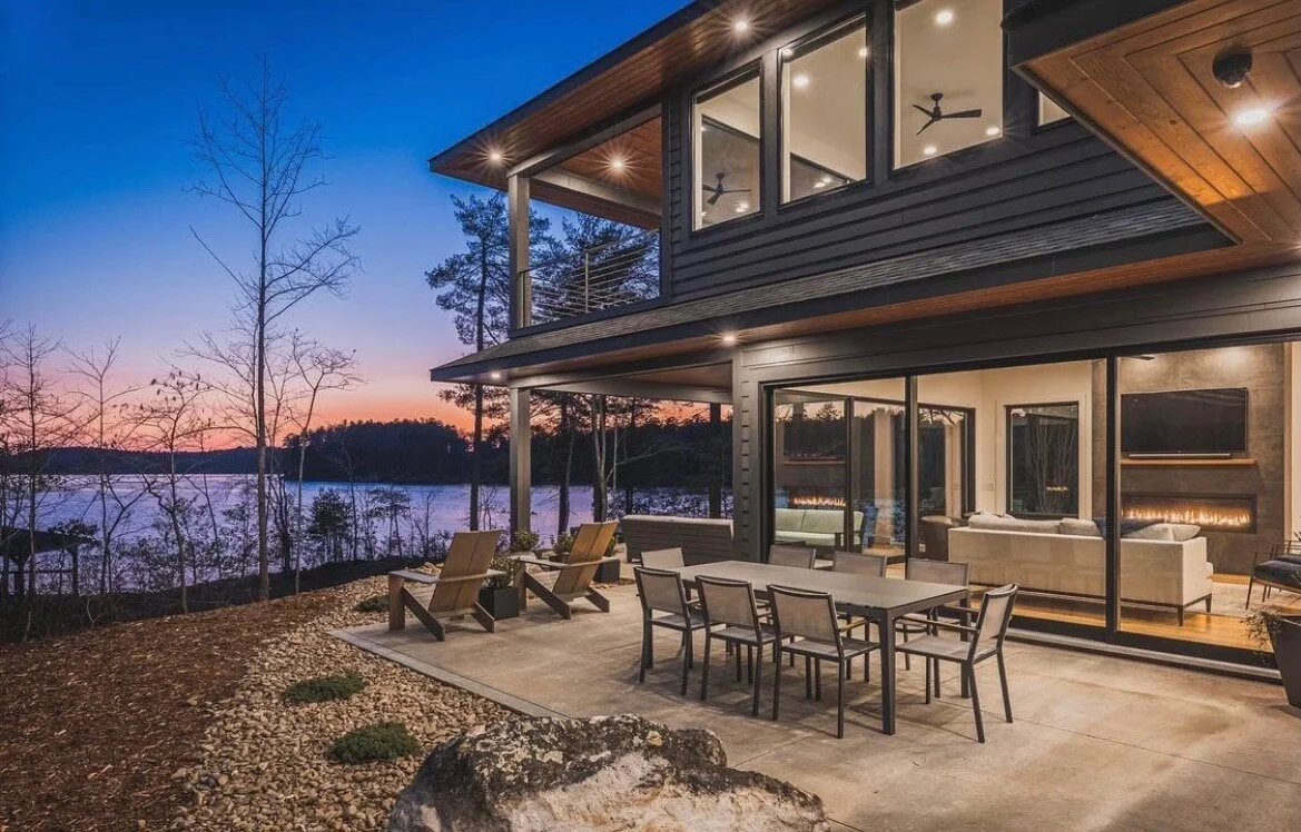 Modern two-story house with a large outdoor patio overlooking a lake during sunset. The patio has outdoor chairs, a dining table, and interior lighting visible through large glass doors and windows.