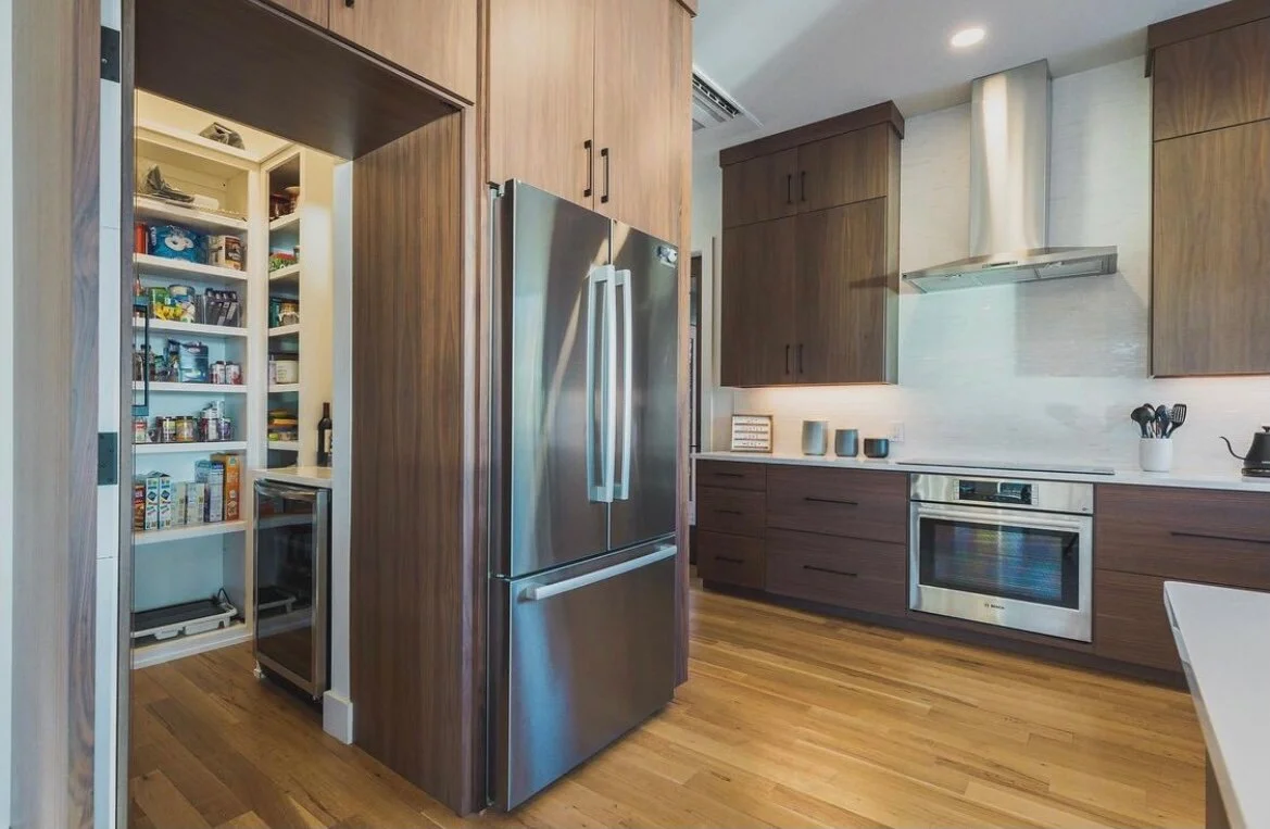 Modern kitchen with wooden cabinets, stainless steel refrigerator, oven, range hood, and open pantry with snacks and food items