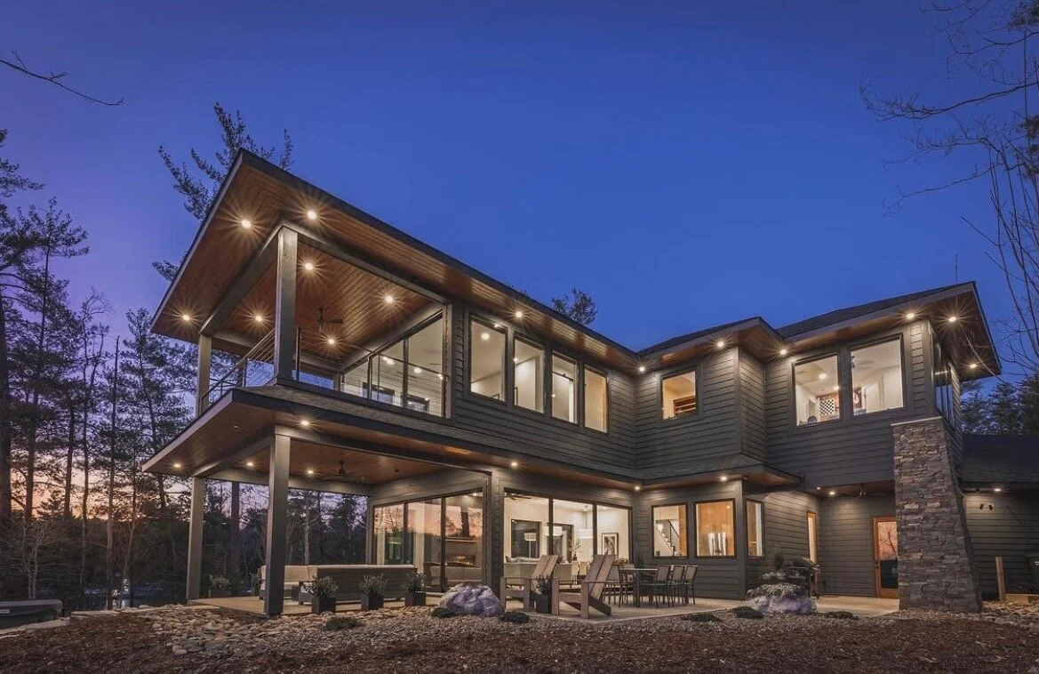 Modern two-story house with large windows, exterior lighting, and outdoor seating area surrounded by trees at dusk.