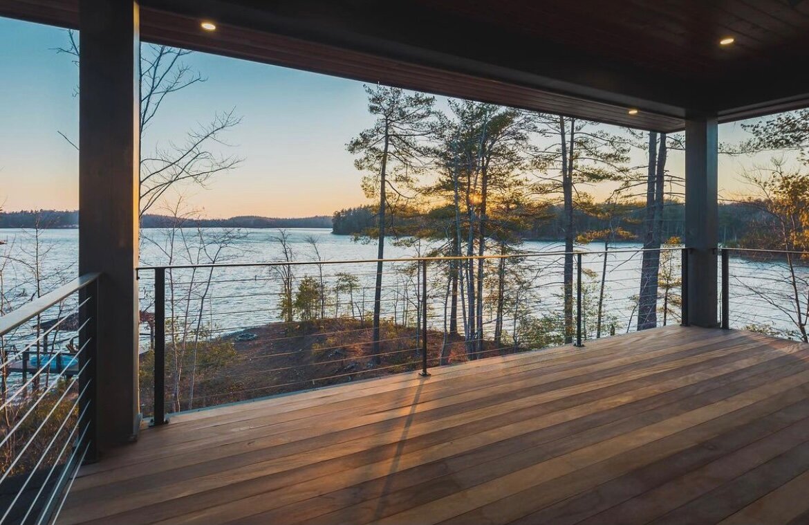 View of a river at sunset from a balcony with wooden flooring and a metal railing, surrounded by trees.