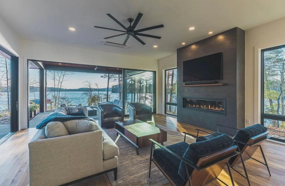 Living room with large windows overlooking a lake, furnished with a beige sofa, black armchairs, a wooden coffee table, and a tile fireplace with a mounted flat-screen TV.