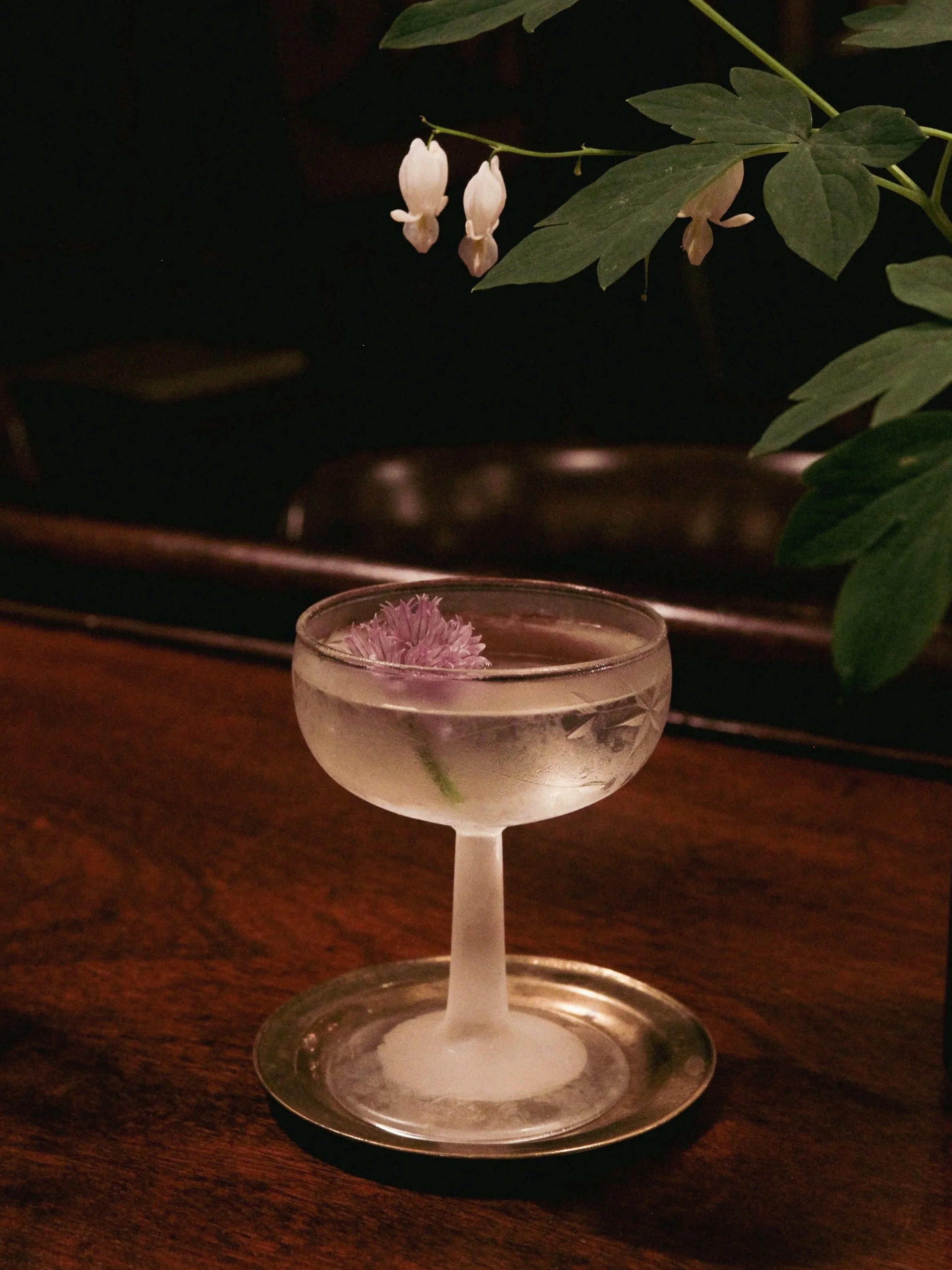 A cocktail glass with a pink flower inside, placed on a wooden surface with greenery and white flowers in the background.