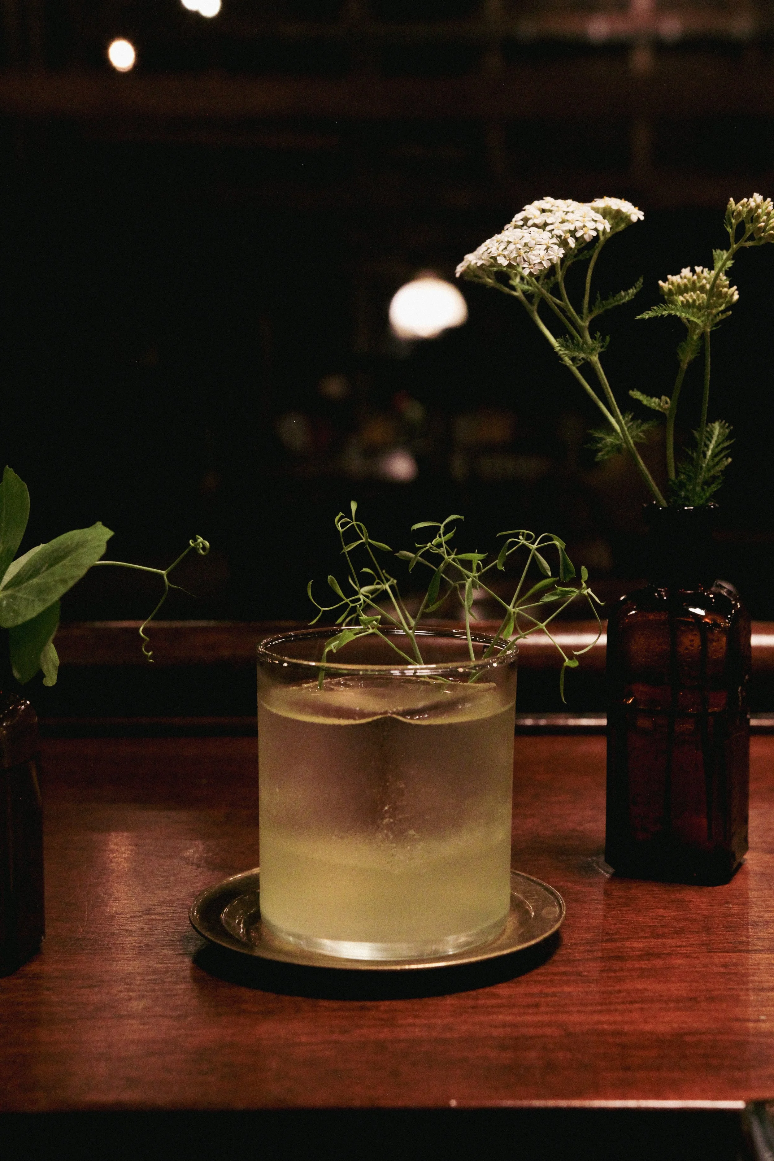 A glass of cocktail with floating greenery on a wooden table, flanked by two small brown vases with white and green flowers, inside a dimly lit room.