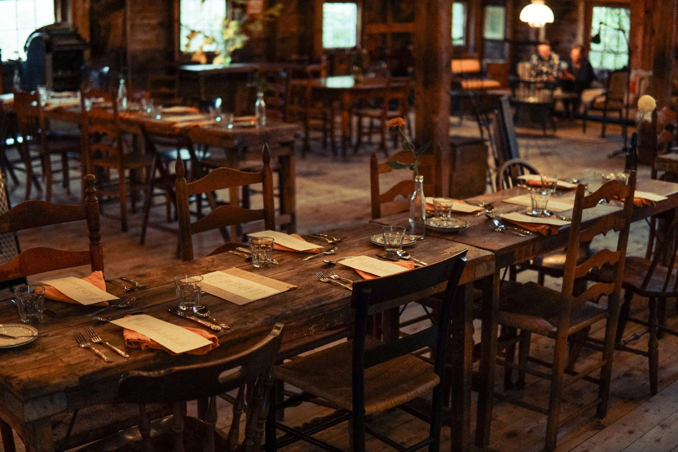 Empty rustic restaurant dining area with wooden tables, chairs, and place settings, including menus, glasses, and napkins, with some guests in the background.