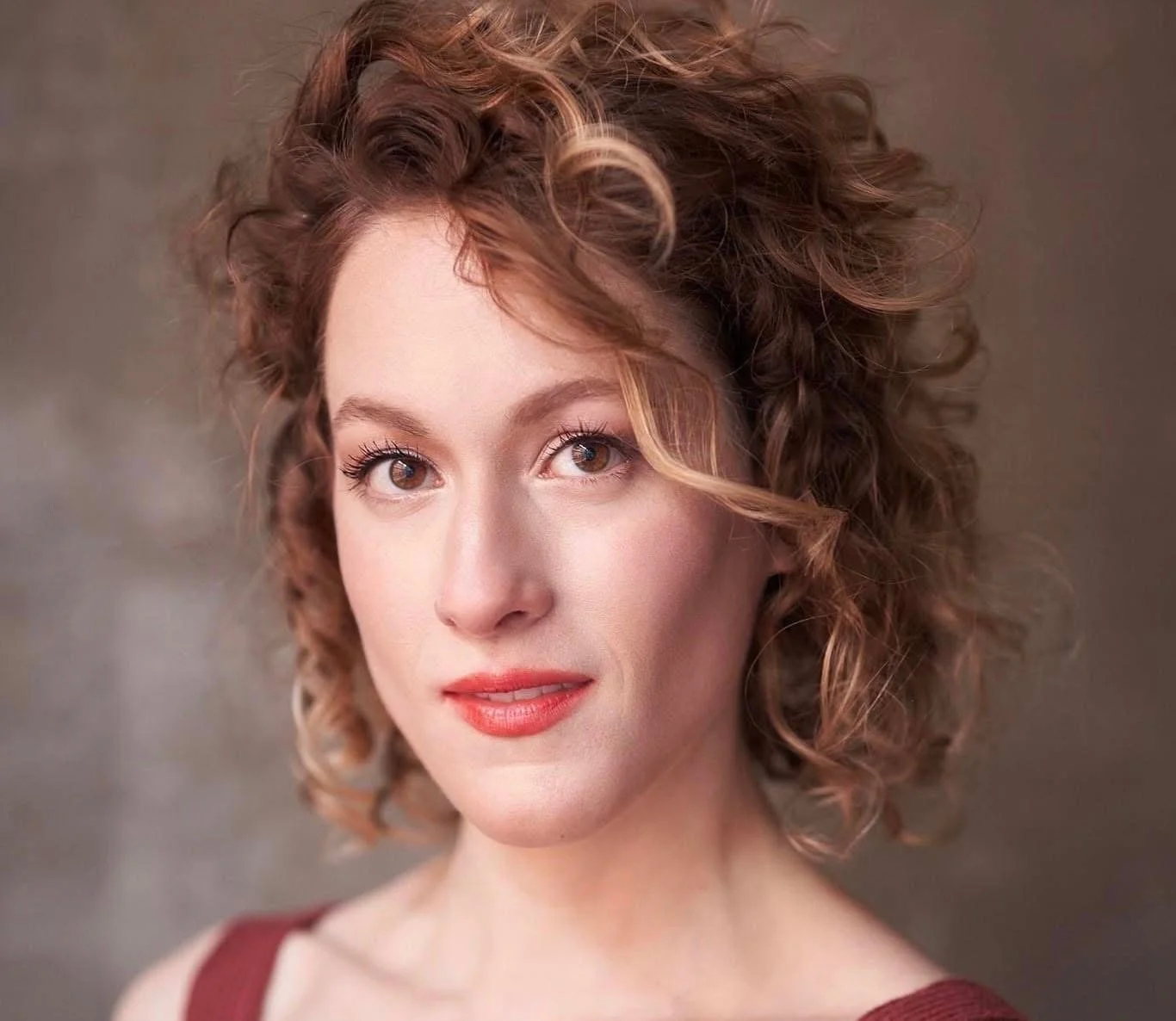 Close-up portrait of a woman with curly red hair and brown eyes, wearing a maroon top, looking at the camera, with a neutral background.