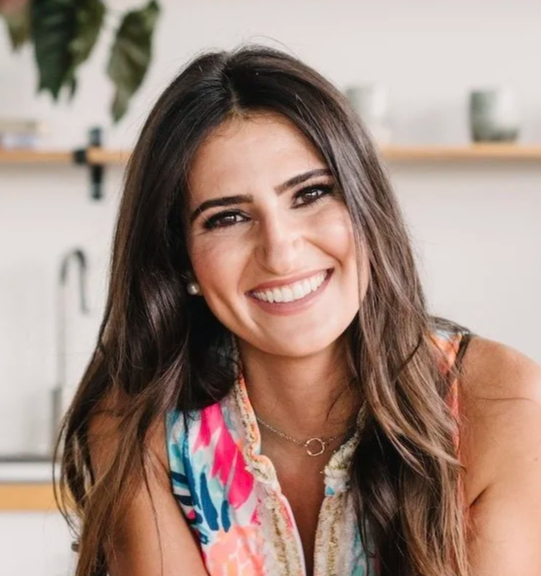 Young woman with long brown hair and a bright smile, wearing a colorful sleeveless top, in a modern kitchen setting.
