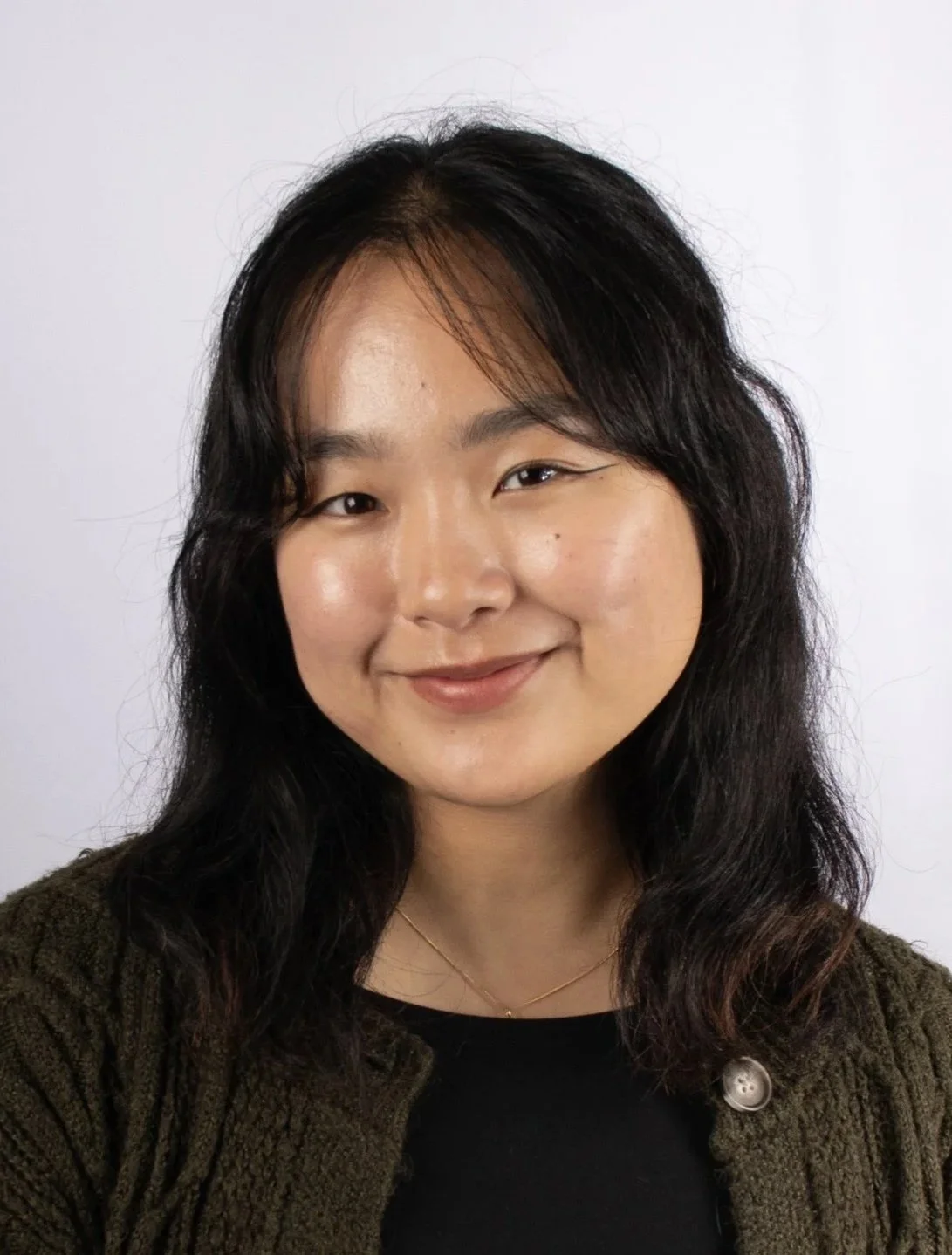 A woman with medium skin, black wavy hair, and a gentle smile, wearing a black top, a green textured cardigan, and a delicate gold necklace, standing against a plain light background.