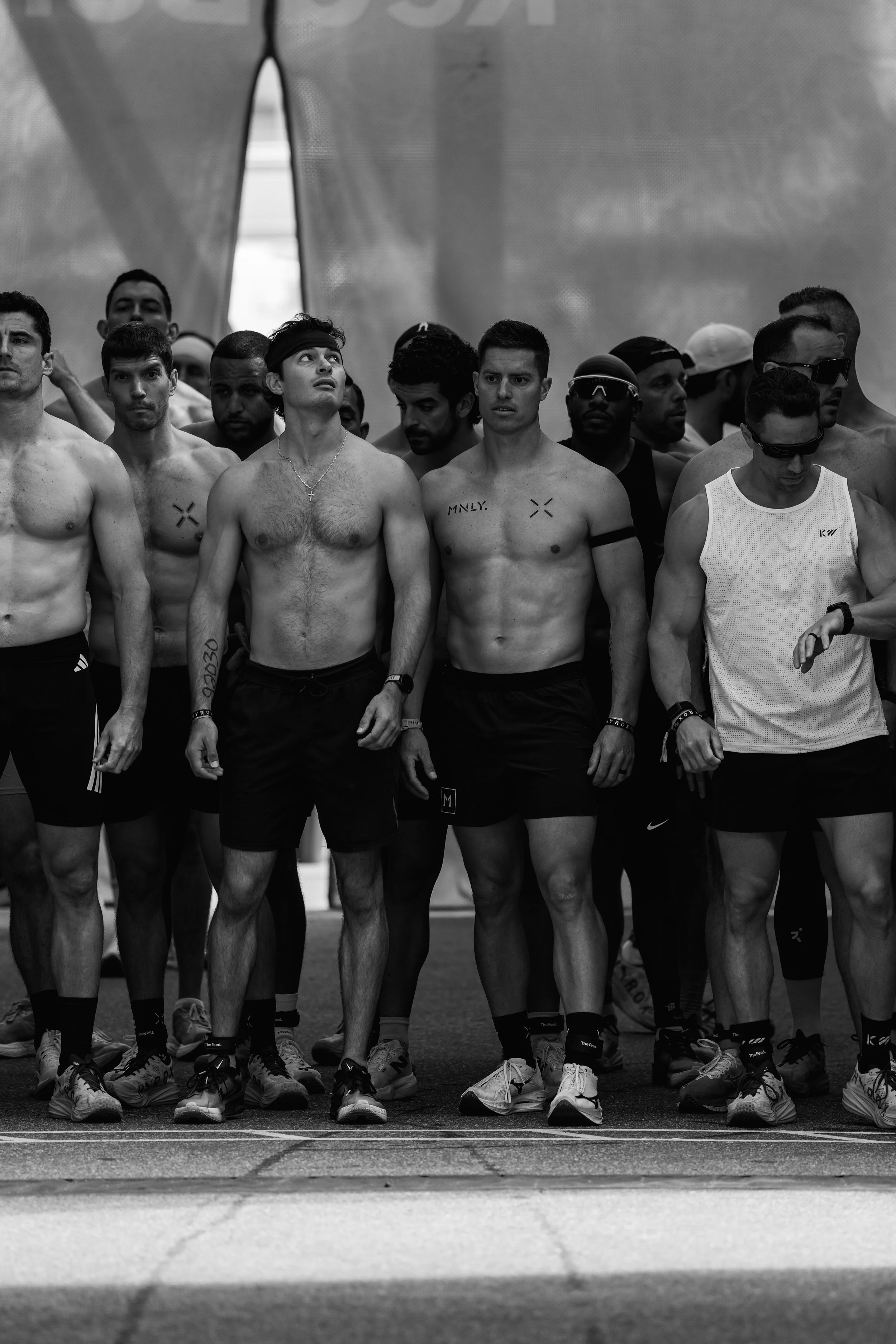 A group of shirtless male athletes standing together at the start line of a race, wearing athletic shoes and shorts, ready to compete in a track event.