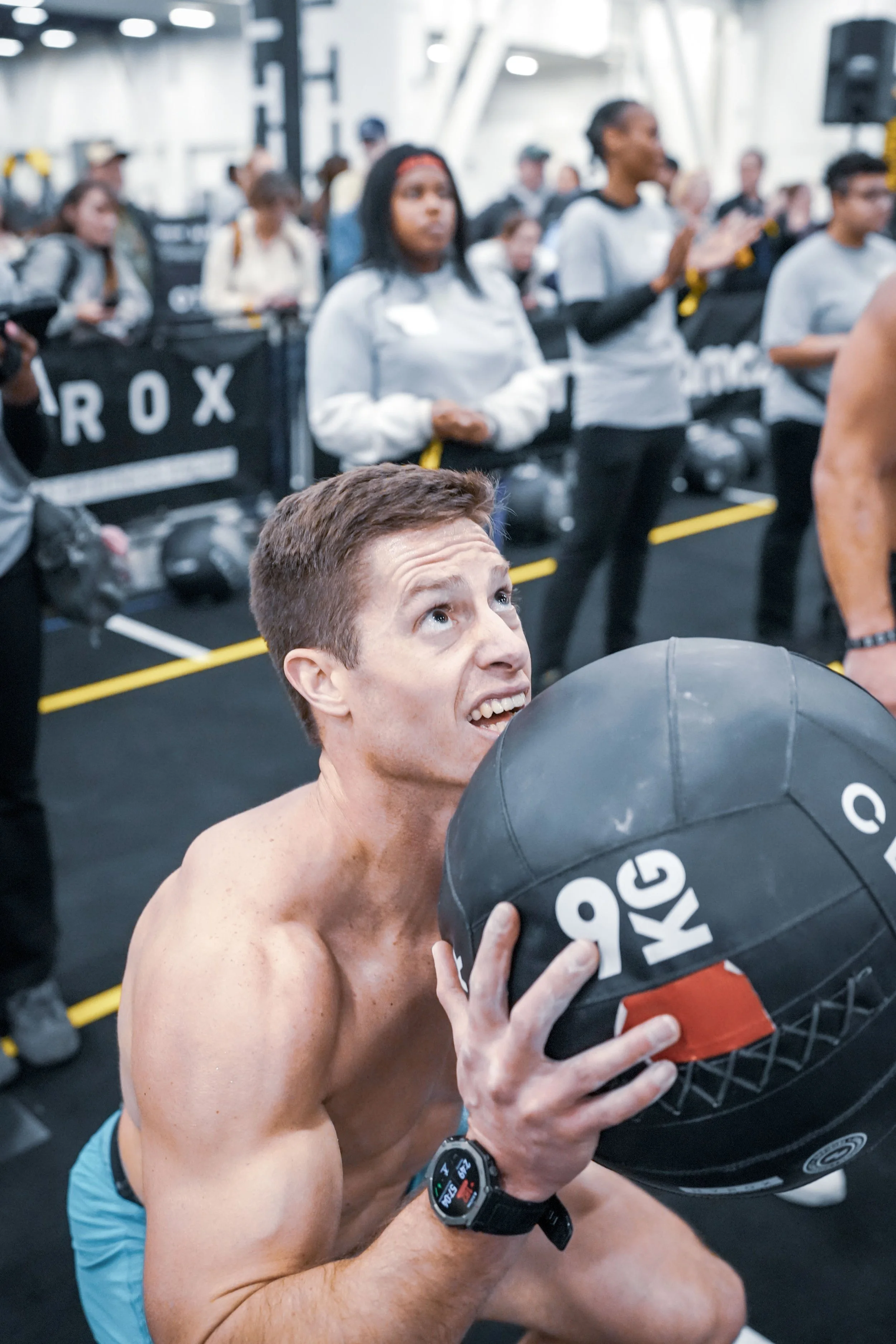 A muscular young man with short brown hair, shirtless, holding a black and orange kettlebell close to his face in a gym. In the background, several women are standing and clapping, dressed in athletic attire. The gym has bright lighting and a visible