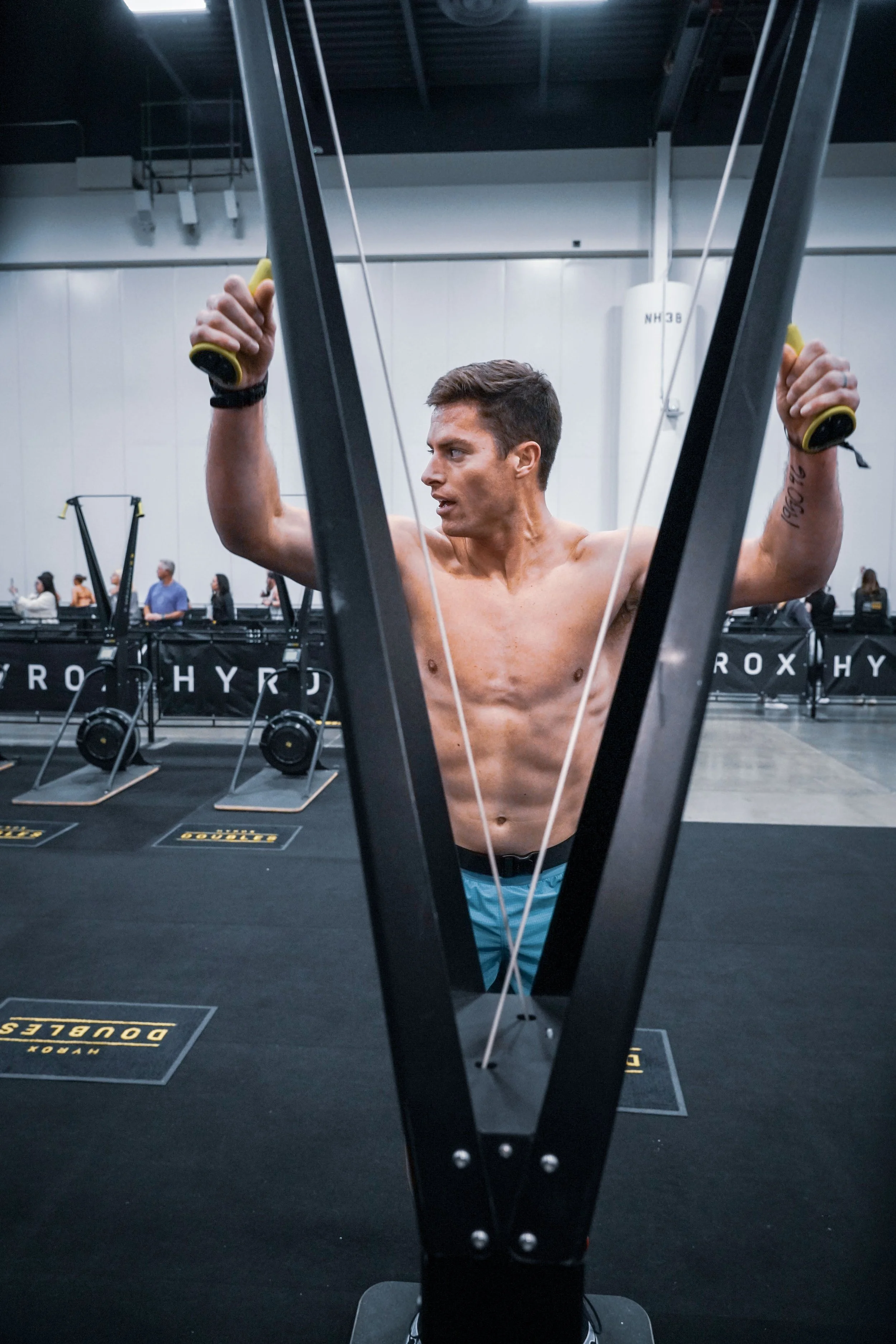 A shirtless man with short brown hair is using an elliptical machine in a gym, with other gym equipment and people visible in the background.