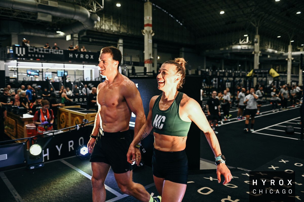 A muscular man and a smiling woman, both wearing athletic clothing, walking together inside a busy CrossFit gym with many people exercising in the background. The gym has high ceilings, industrial lighting, and signage that reads 'HYROX CHICAGO.'