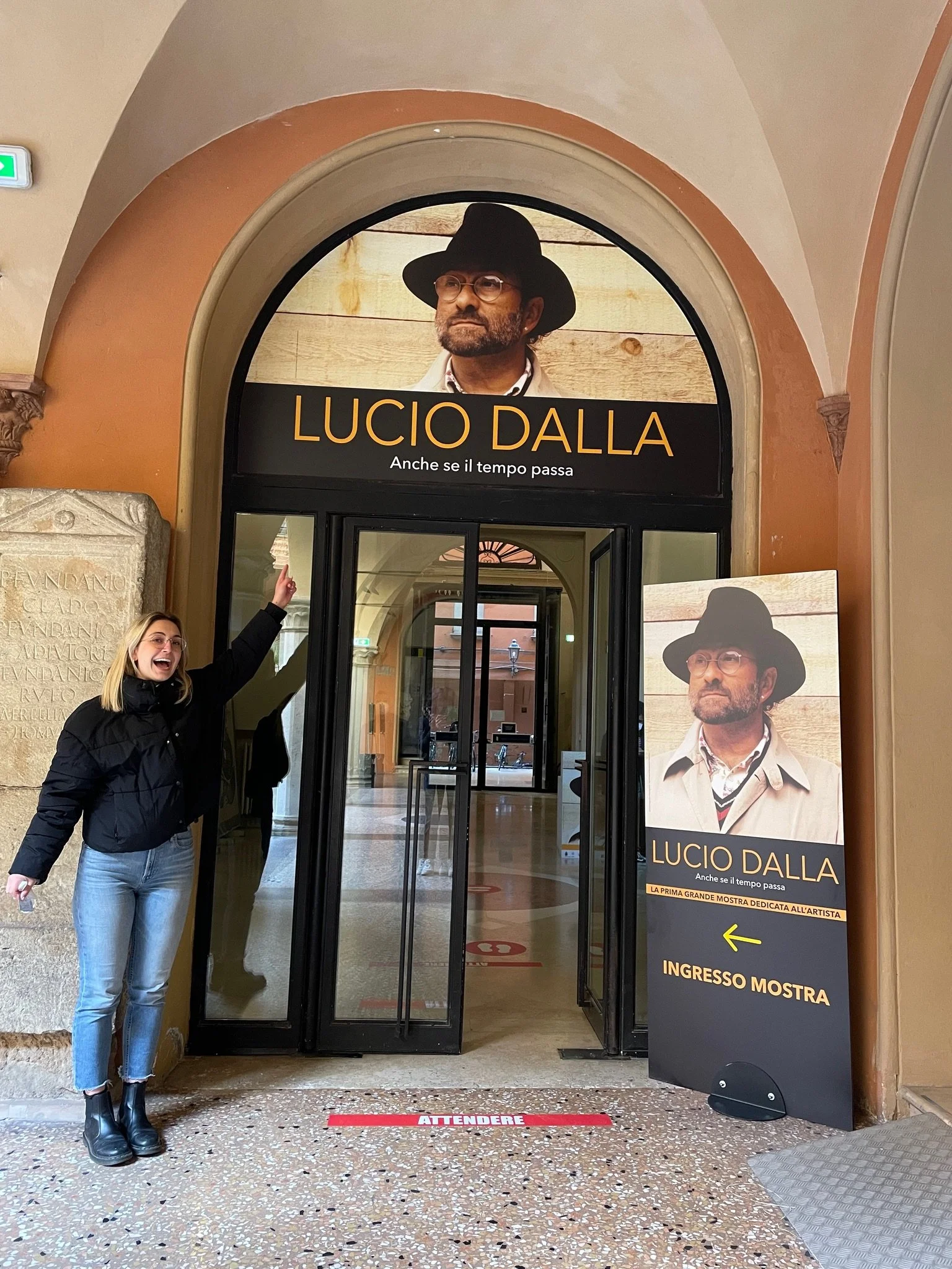 A woman in a black jacket and blue jeans is smiling and pointing at a large poster of Lucio Dalla outside an exhibition entrance. The entrance has a large arched marquee with a photograph of Lucio Dalla wearing a hat and glasses. There is a smaller promotional sign on a stand to the right of the entrance with text in Italian. The woman is standing on a terrazzo floor with a red waiting line marker that says "ATTENDERE."