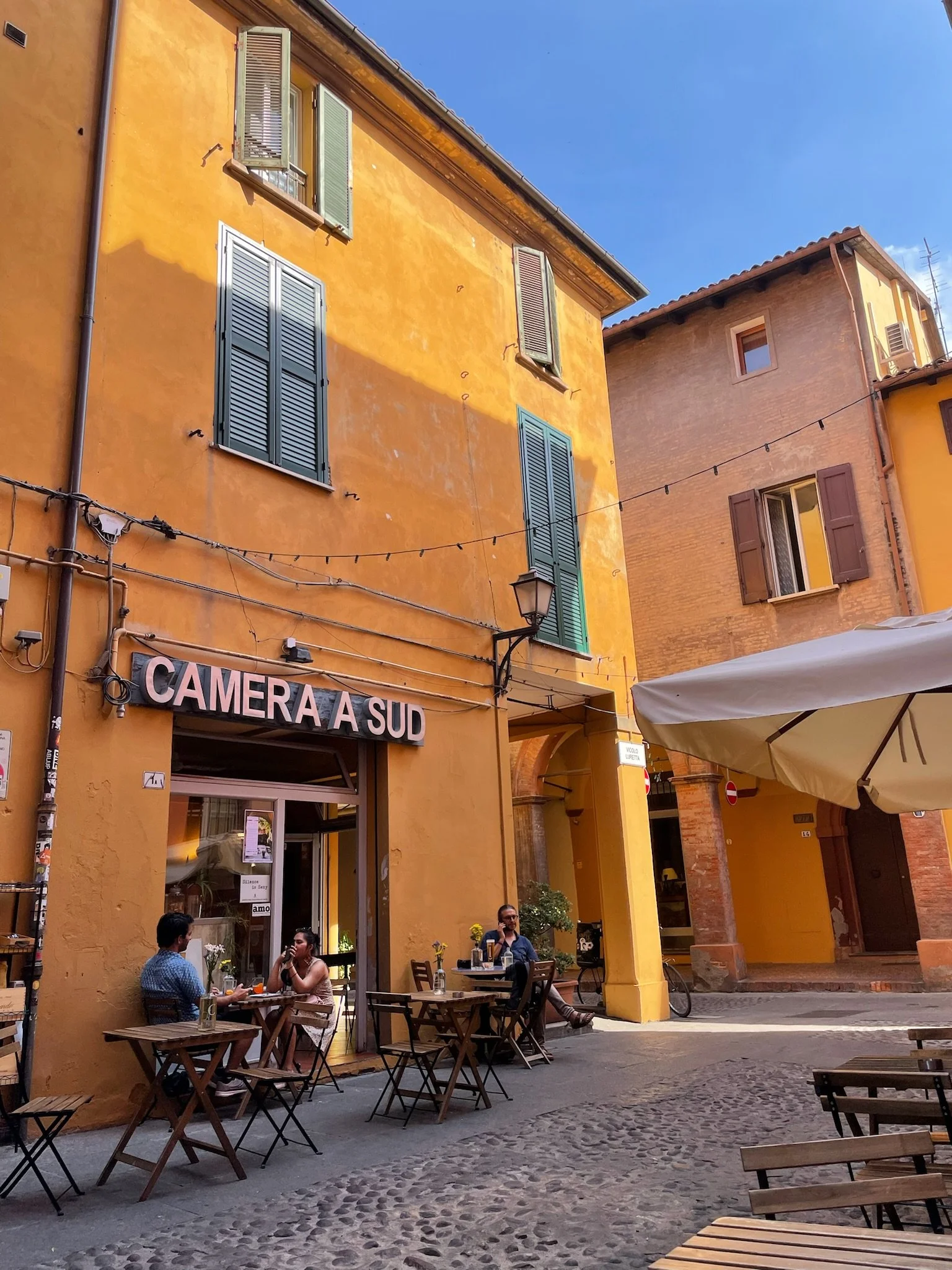 People sitting at outdoor tables of a café called 'Camera A Sud' on a cobblestone street, surrounded by yellow buildings with green and brown shutters, under a clear blue sky.