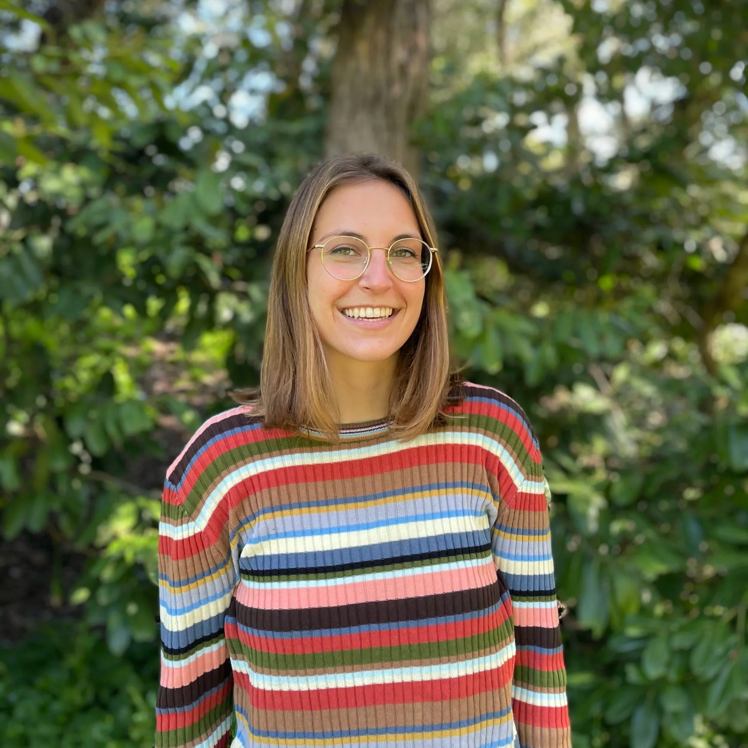 A woman with shoulder-length brown hair, wearing glasses and a multicolored striped sweater, smiling outdoors in front of green foliage.