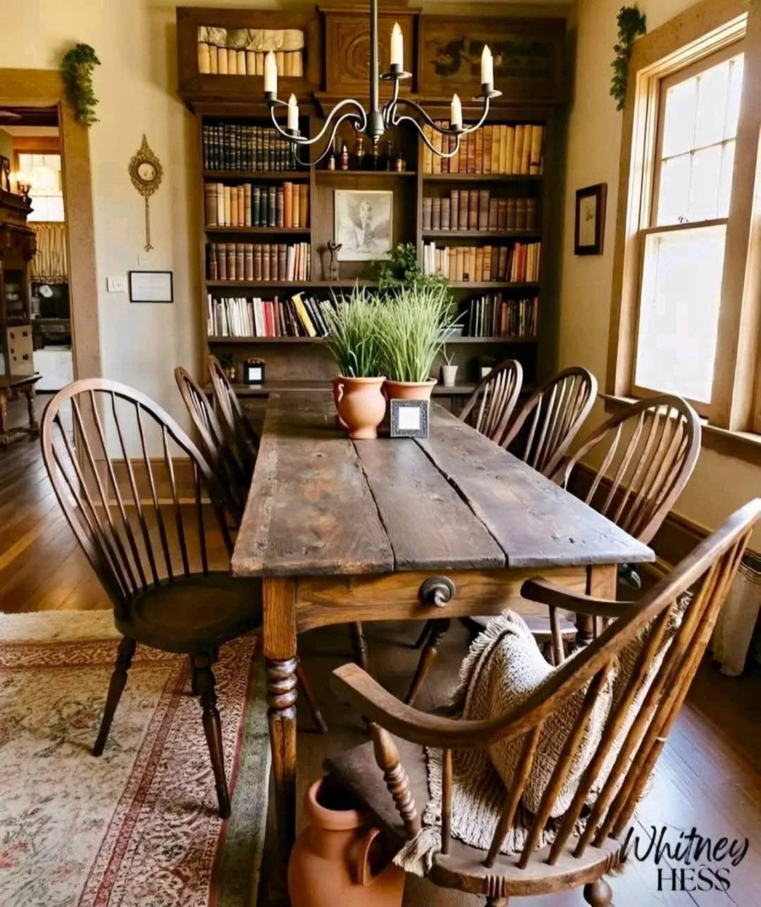 A rustic wooden dining table with eight wooden chairs, two potted plants, and a small picture frame, set in a cozy dining room with bookshelves, framed pictures, a chandelier, window, and wooden floors.