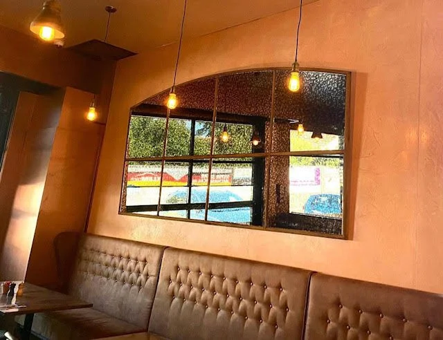 Interior of a cafe with a tufted booth, hanging Edison-style light bulbs, and a large vintage mirror reflecting outside view.