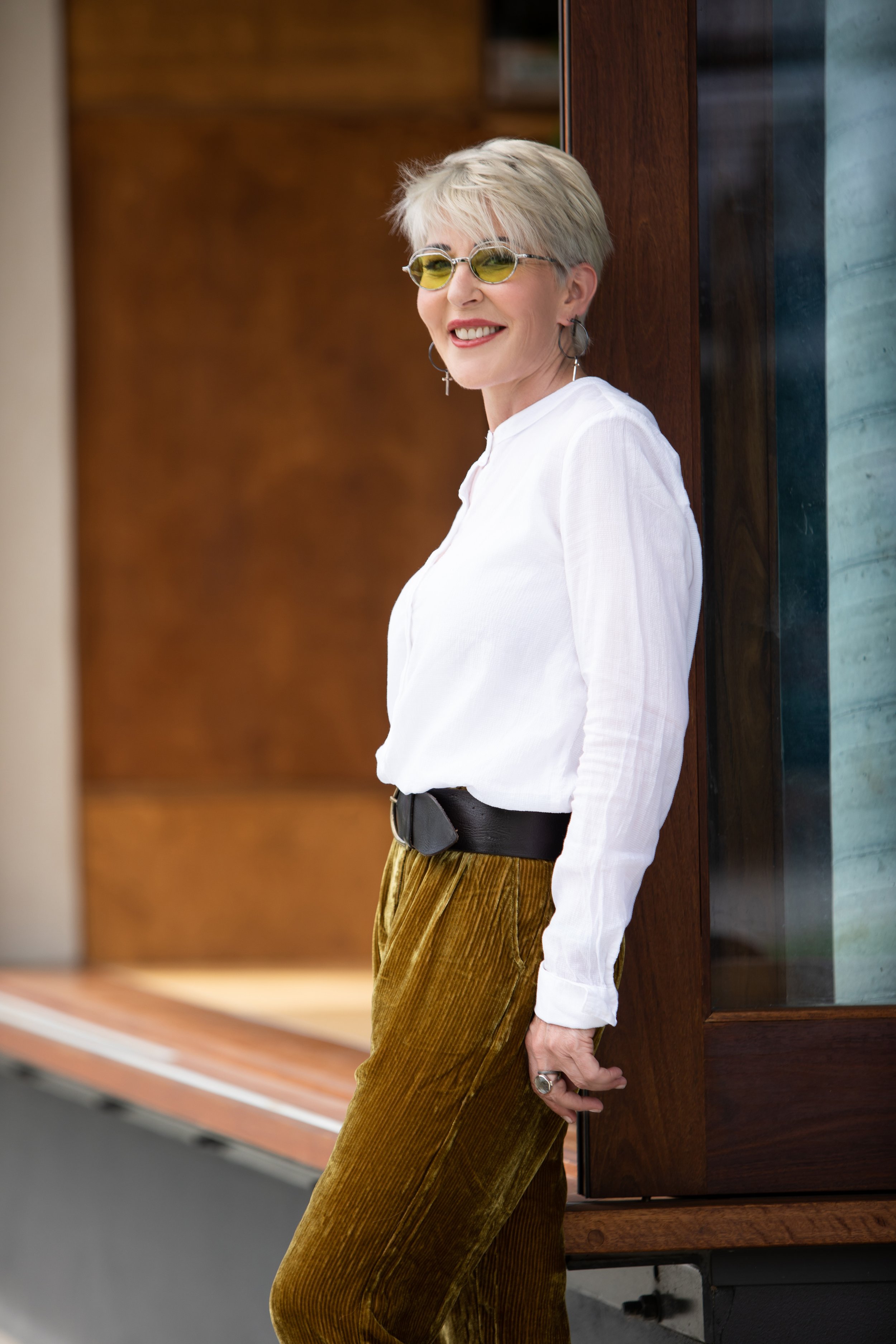 A woman with short, styled blonde hair wearing yellow sunglasses, a white long sleeve top, gold velvet pants, and jewelry, leaning against a wooden wall outside a building and smiling.