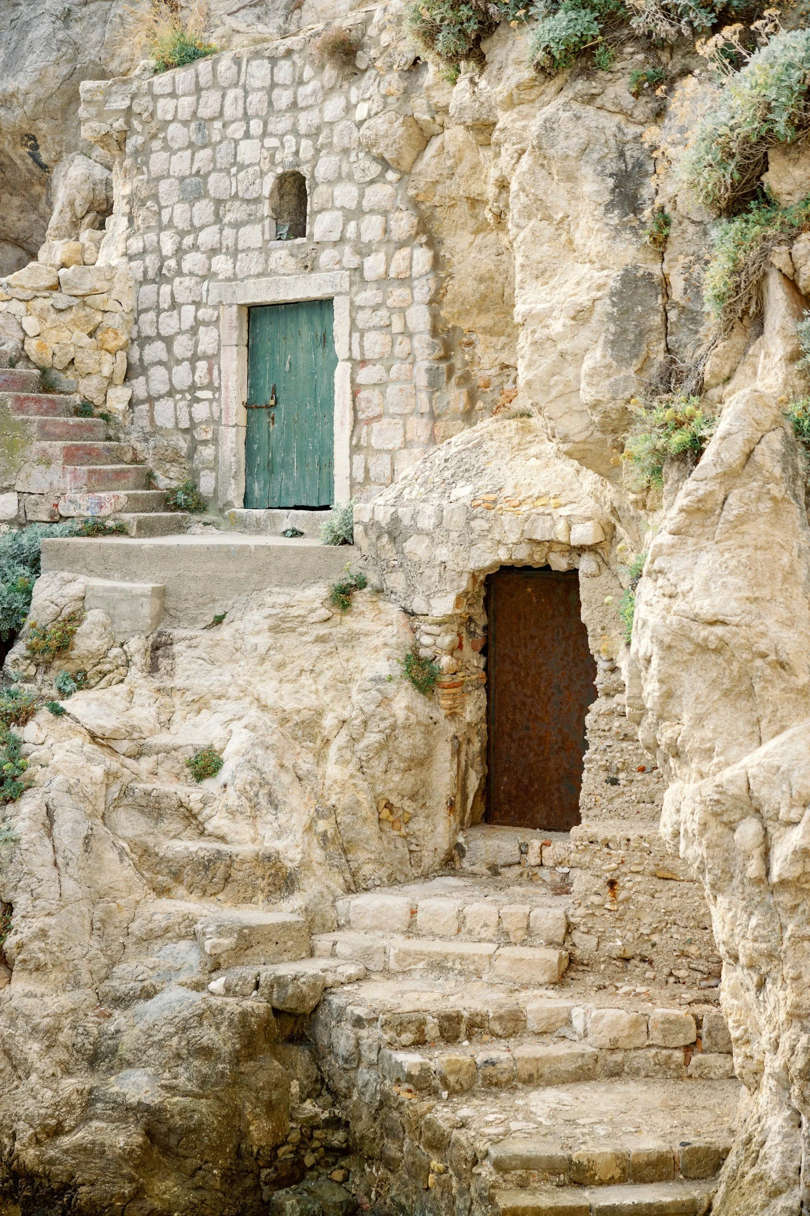 Stone stairs leading up to a small stone structure with a green door and a rusted metal door embedded in a rocky hillside, surrounded by natural stone and sparse vegetation.
