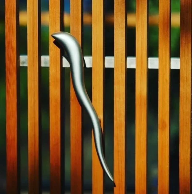 A metallic hook or tool hanging on a wooden slat railing with blurred green and yellow background.