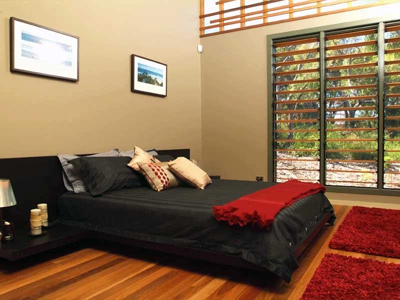 A modern bedroom with a black bed covered in black bedding, several pillows, and a red throw blanket. There are two red area rugs on the wooden floor, a nightstand with candles and a lamp, two framed pictures on the wall, and large windows with wooden slats allowing natural light.