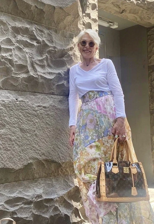 An elderly woman with white hair, wearing sunglasses, a white long-sleeve shirt, light-colored floral pattern pants, and holding a black and tan designer handbag, standing outdoors against a stone wall.