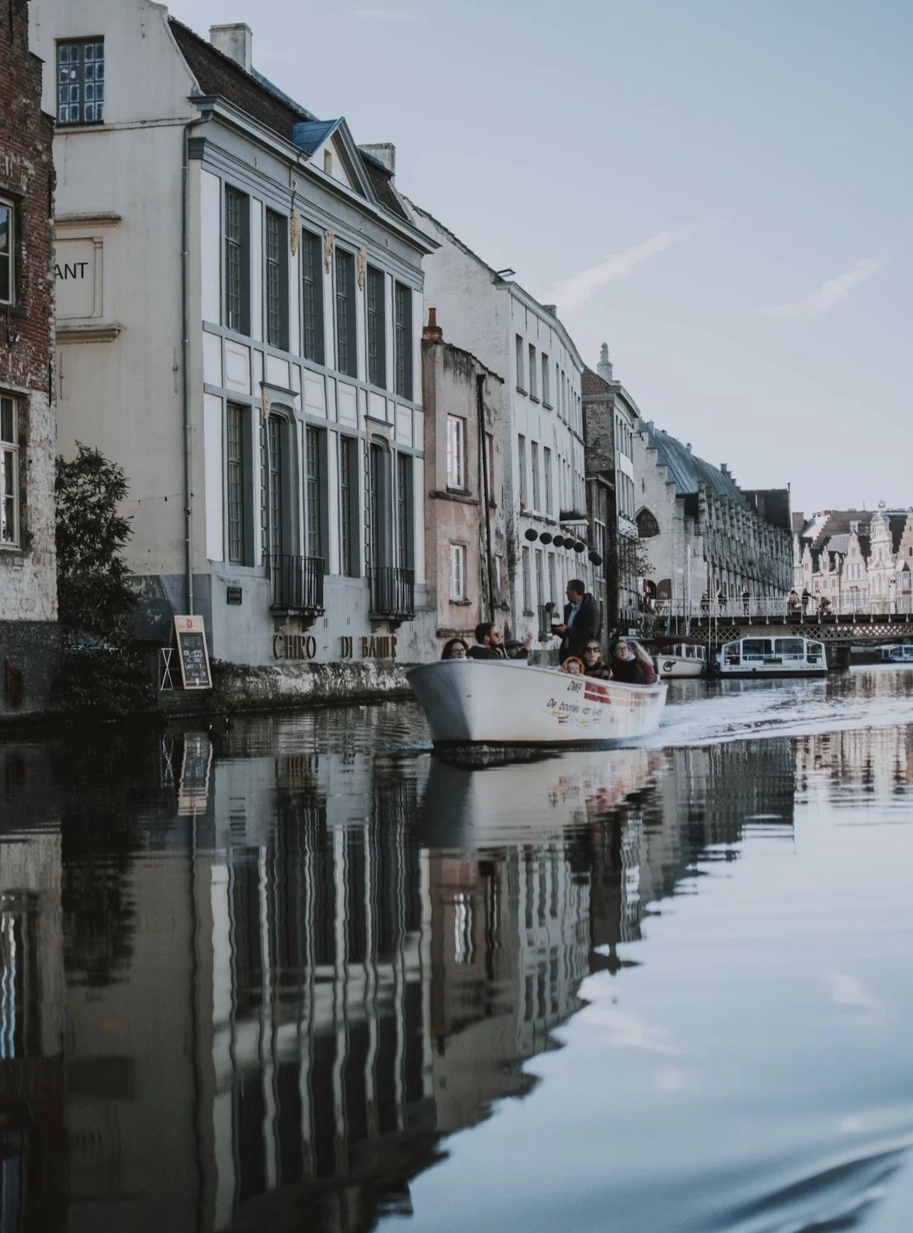 A small boat with passengers on a canal in a European city, with historic buildings and a bridge in the background.