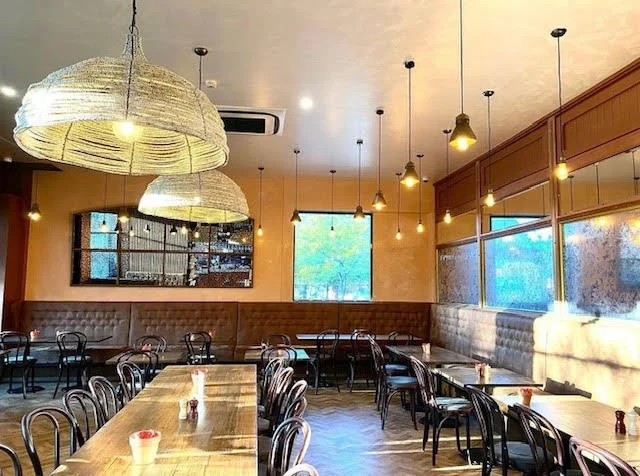 Empty restaurant dining area with wooden tables, black chairs, large woven pendant lights, wall-mounted picture frames, and windows letting in natural light.