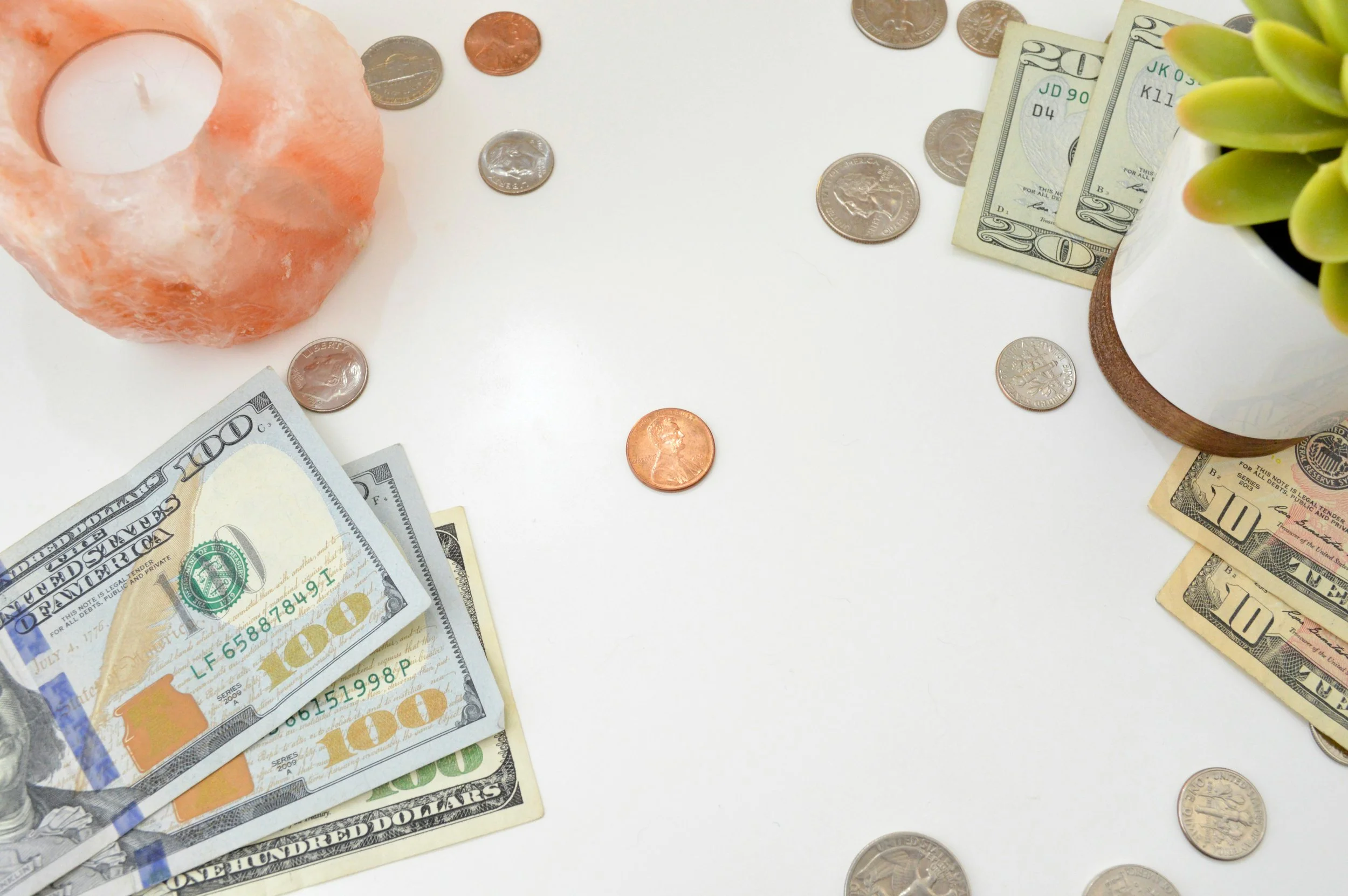 U.S. currency including $100, $20, $10 bills, and various coins on a white surface with a salt candle holder and a potted succulent.
