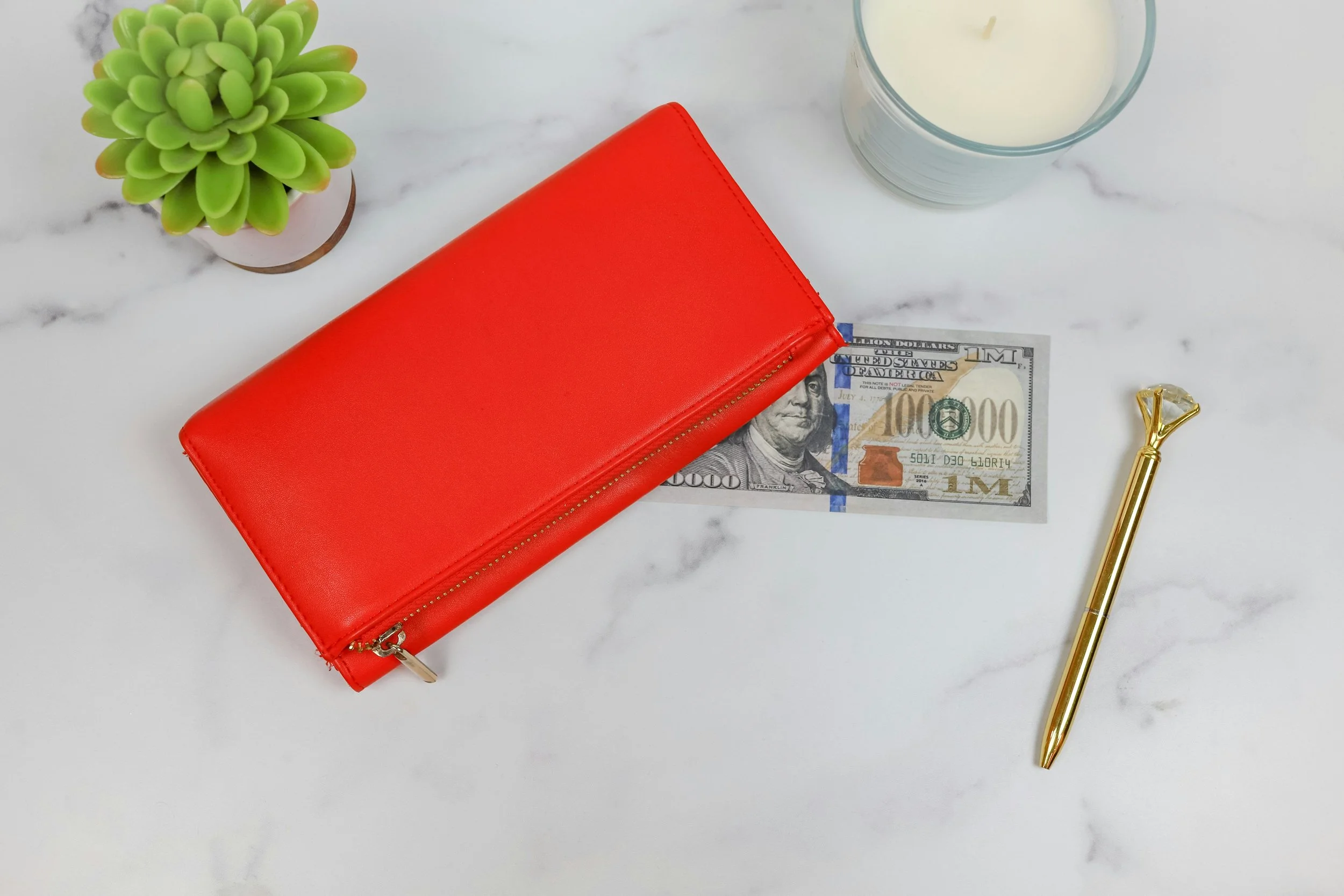 Red wallet, novelty $100,000 bill, green succulent, white candle, gold pen with diamond-shaped top on marble surface.