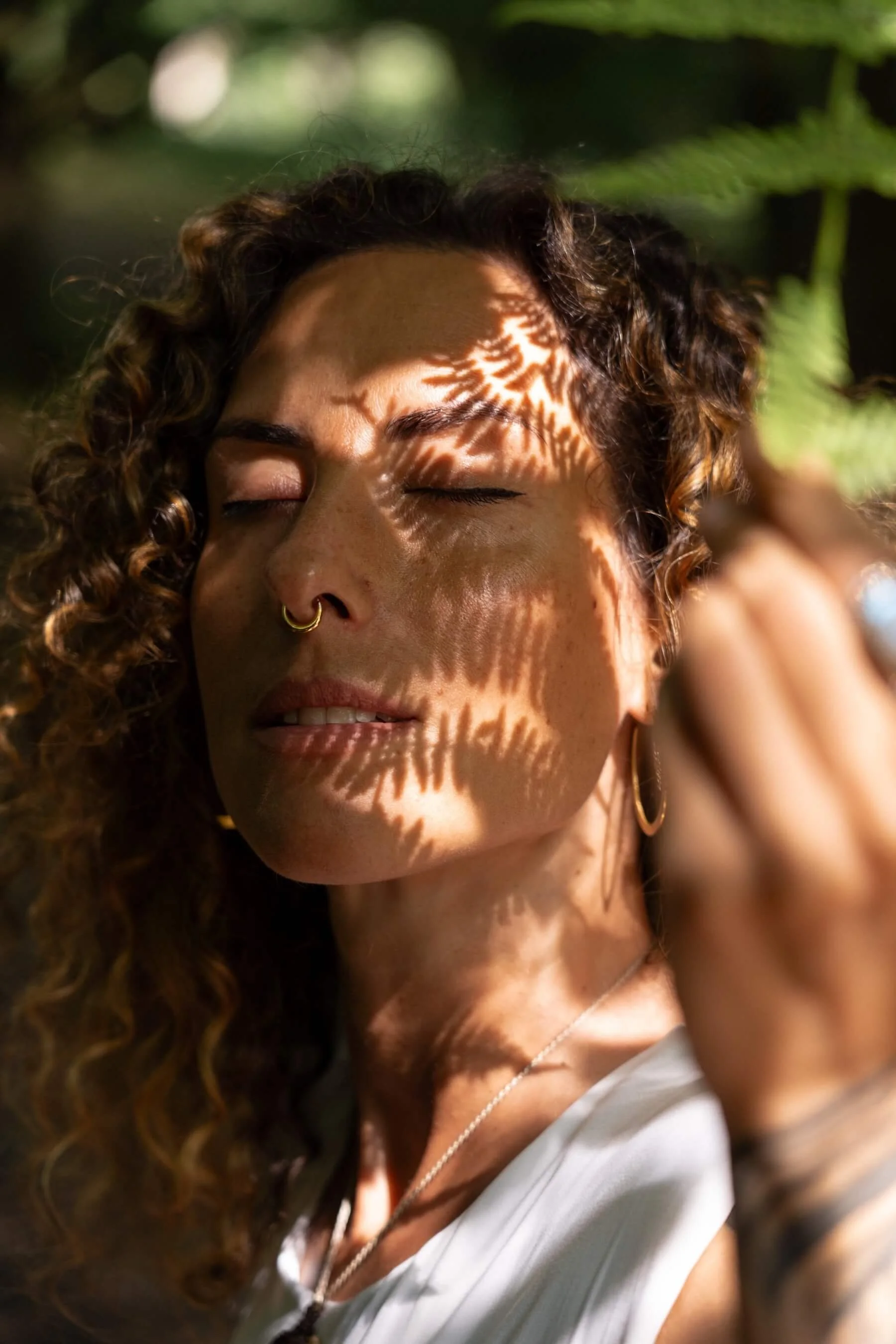 Ana con cabello rizado, maquillaje natural, usando aretes de aro y un collar, con sombra de hojas en su rostro, en un entorno natural y soleado.