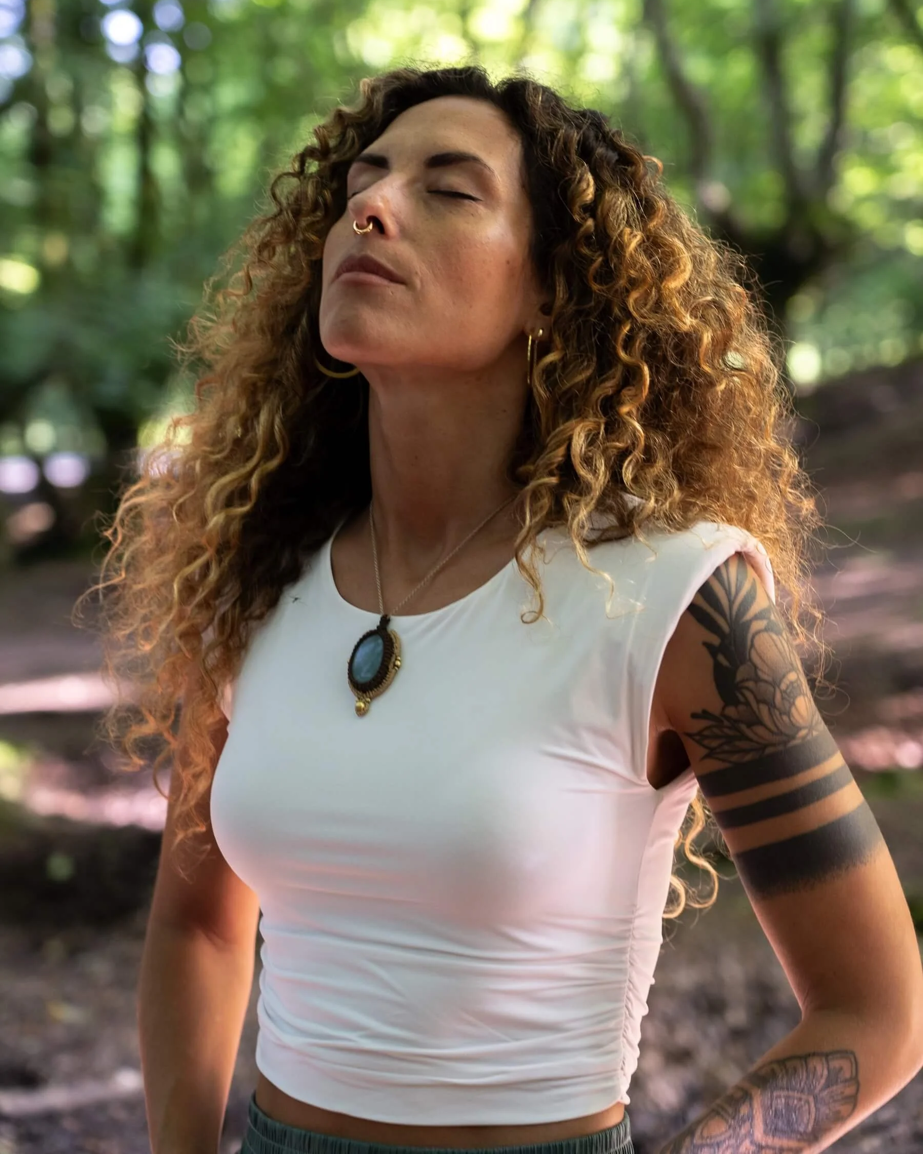 Ana con cabello rizado y tatuajes, con los ojos cerrados, en un bosque con luz solar filtrándose entre los árboles.