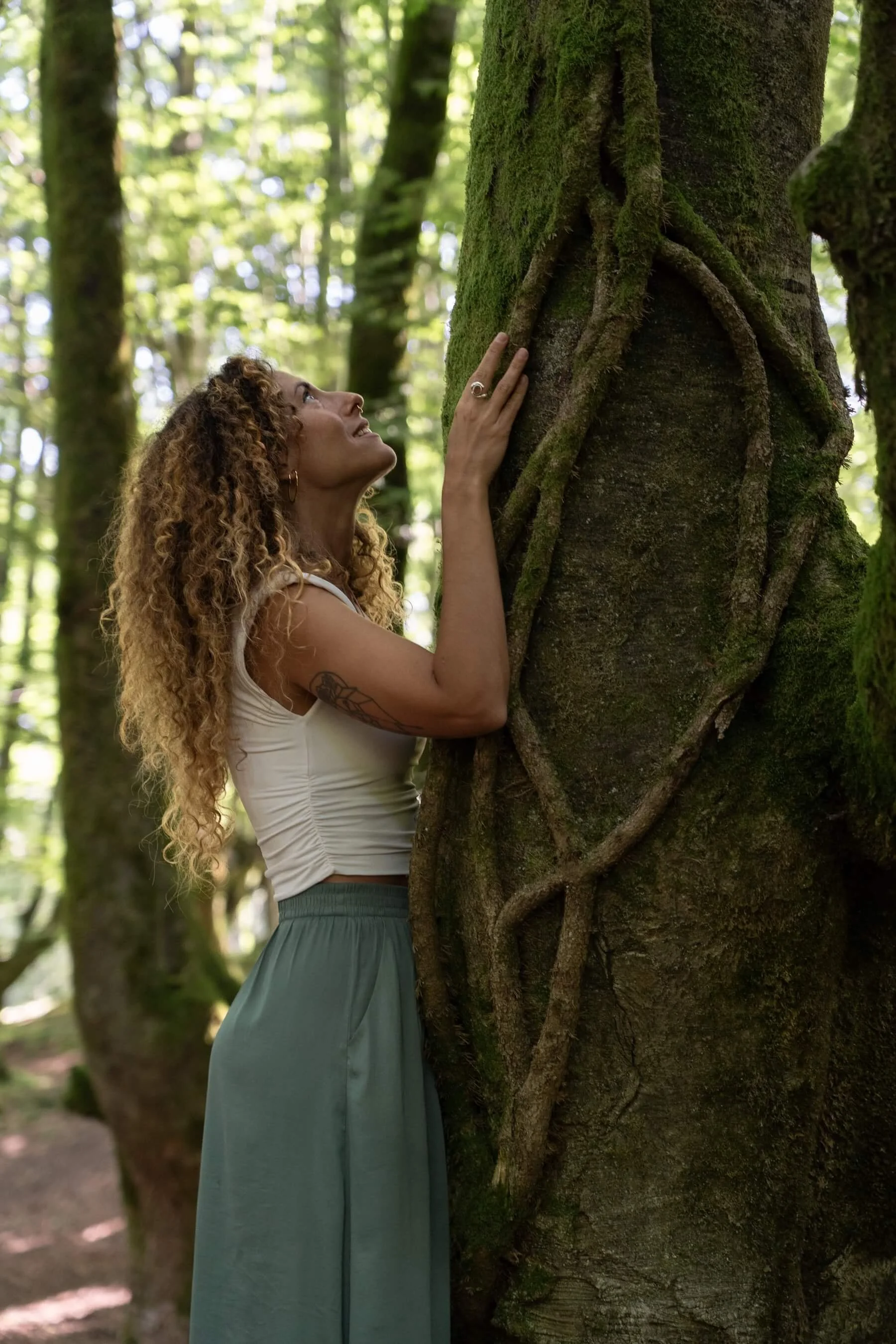 Ana con cabello rizado y tatuajes en el brazo sostiene un árbol en un bosque.