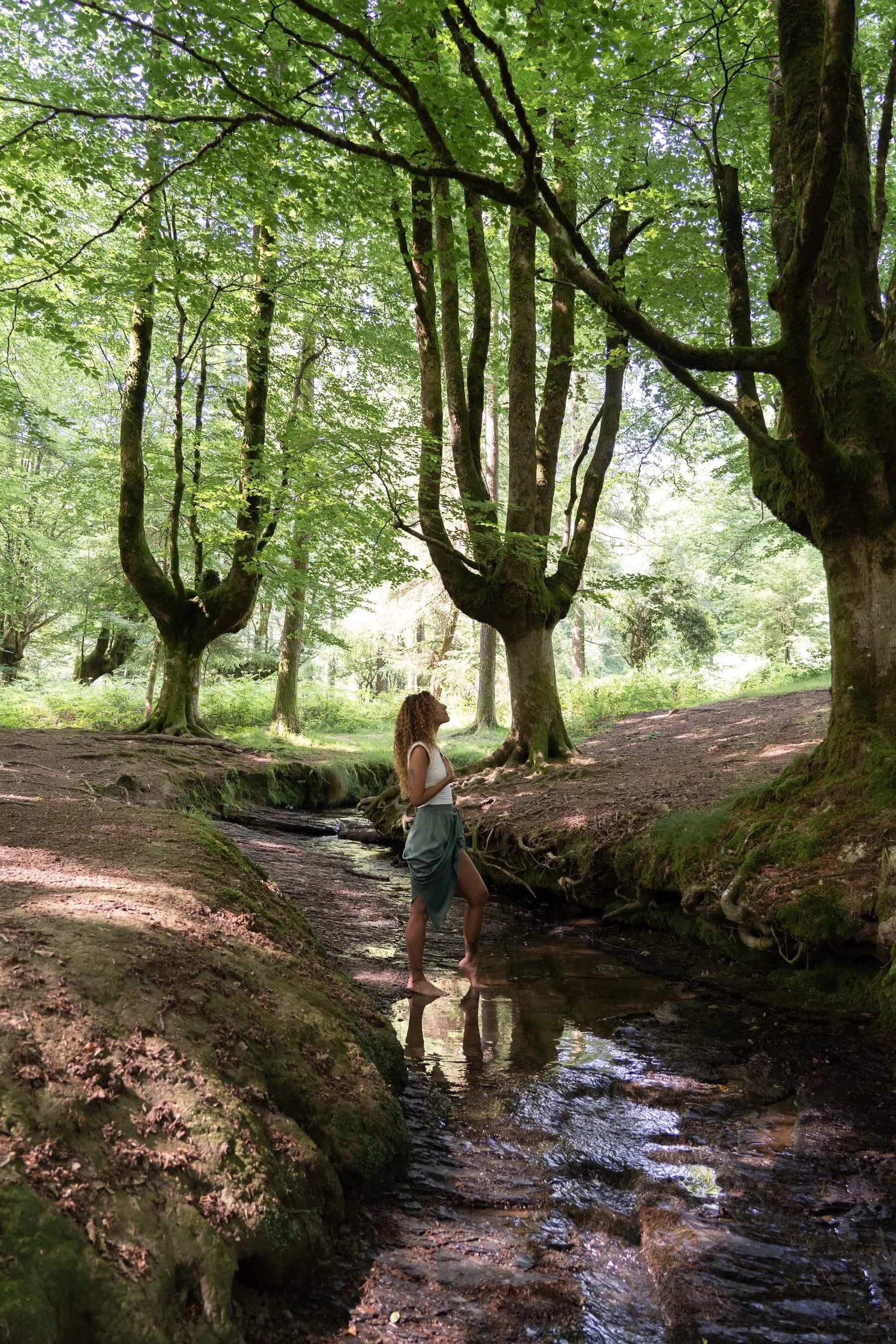 Ana vestida con blusa blanca y falda gris, parada de perfil, de pie en un río rodeado de árboles verdes en un bosque.