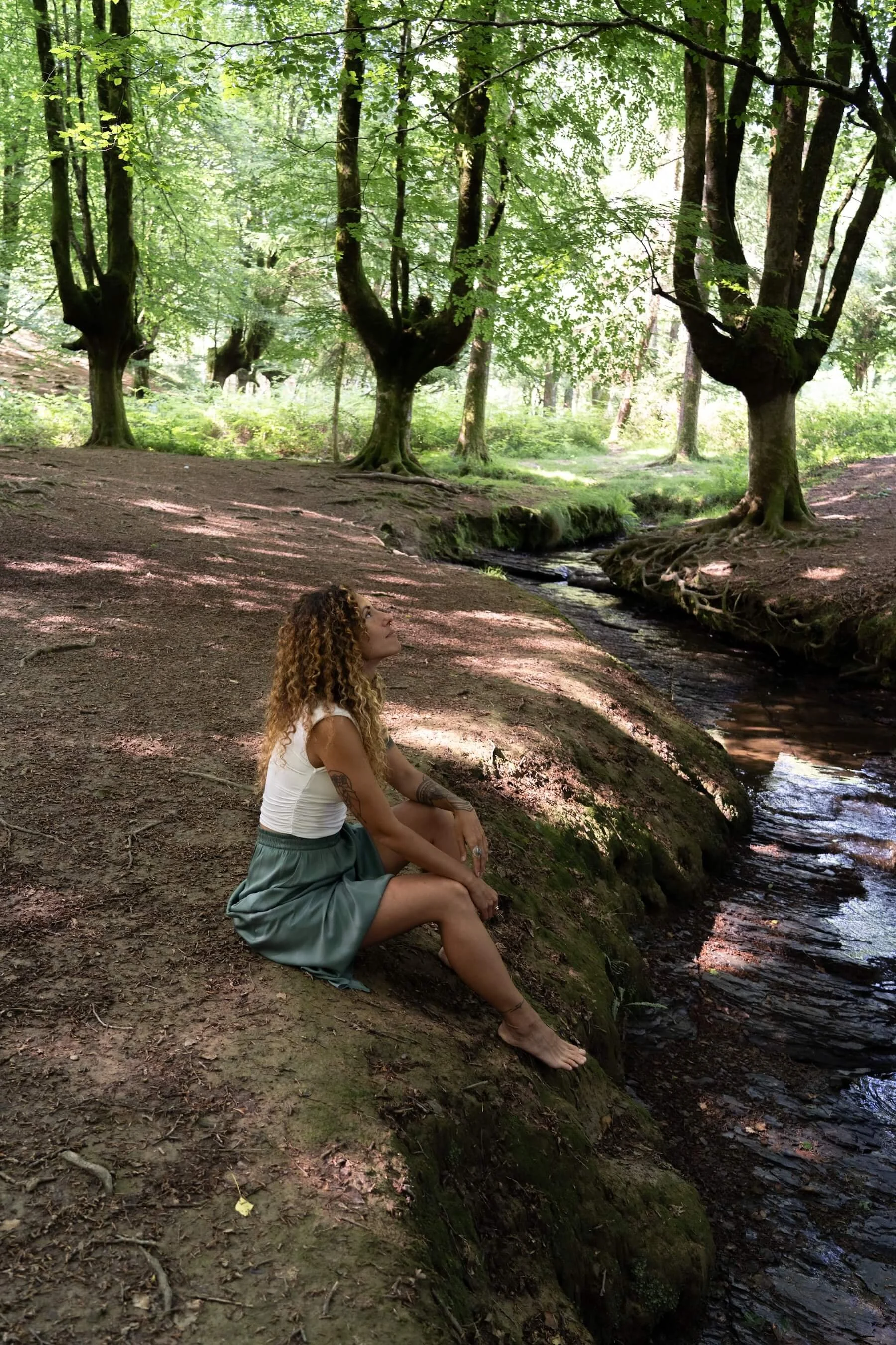 Mujer con cabello rizado sentado en la orilla de un río en un bosque con árboles verdes.