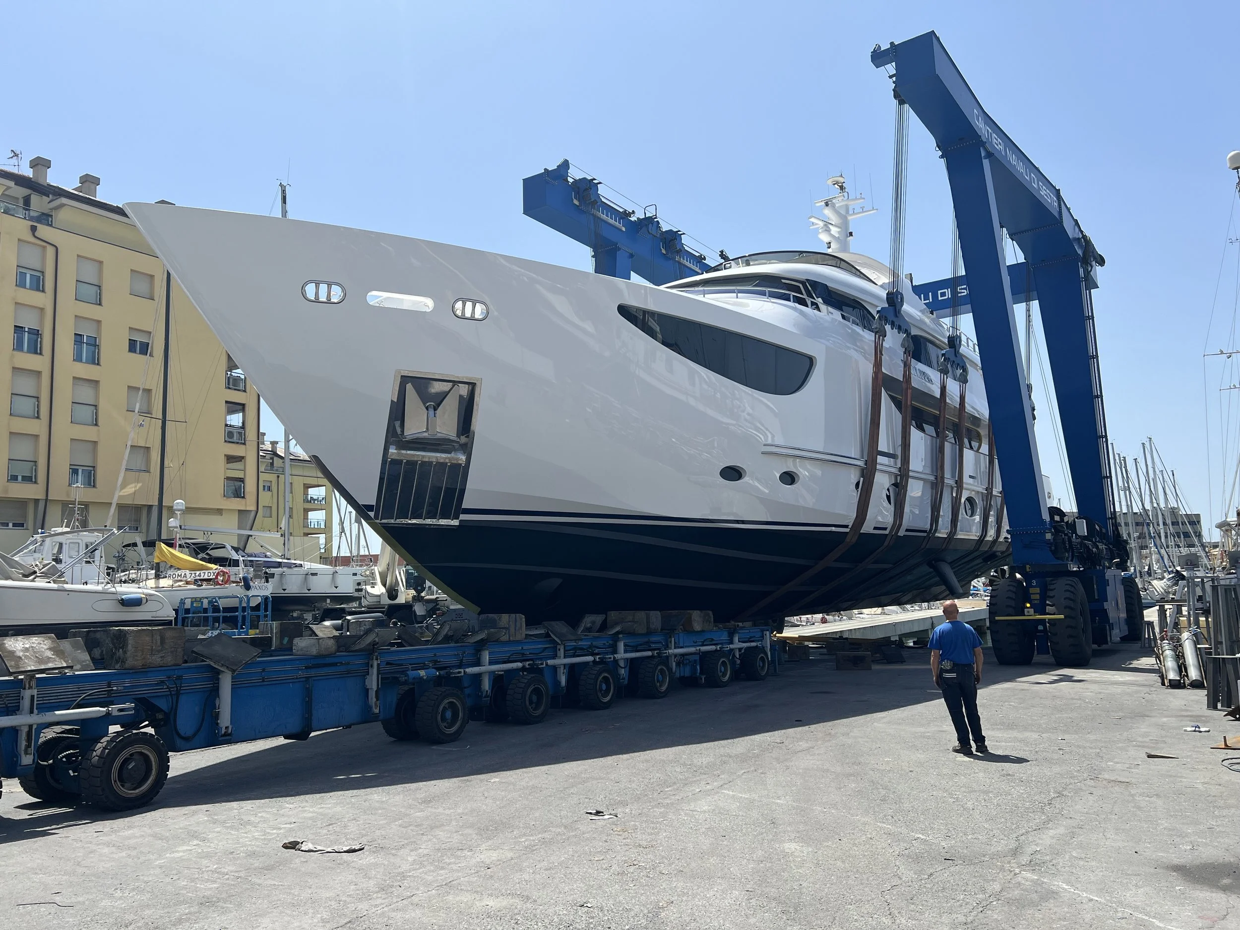 A large yacht is being lifted by a blue crane at a marina, with several smaller boats docked nearby and residential buildings in the background.