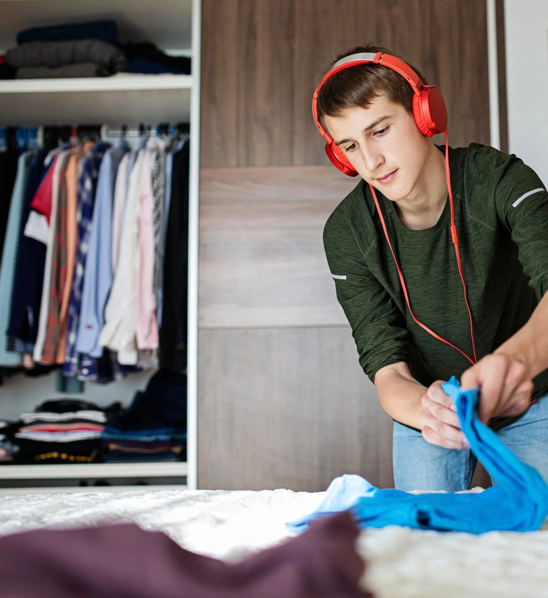 A young man with brown hair wearing red headphones and a dark green shirt sitting on the floor in a room with a closet of clothes in the background, looking down and smiling softly.