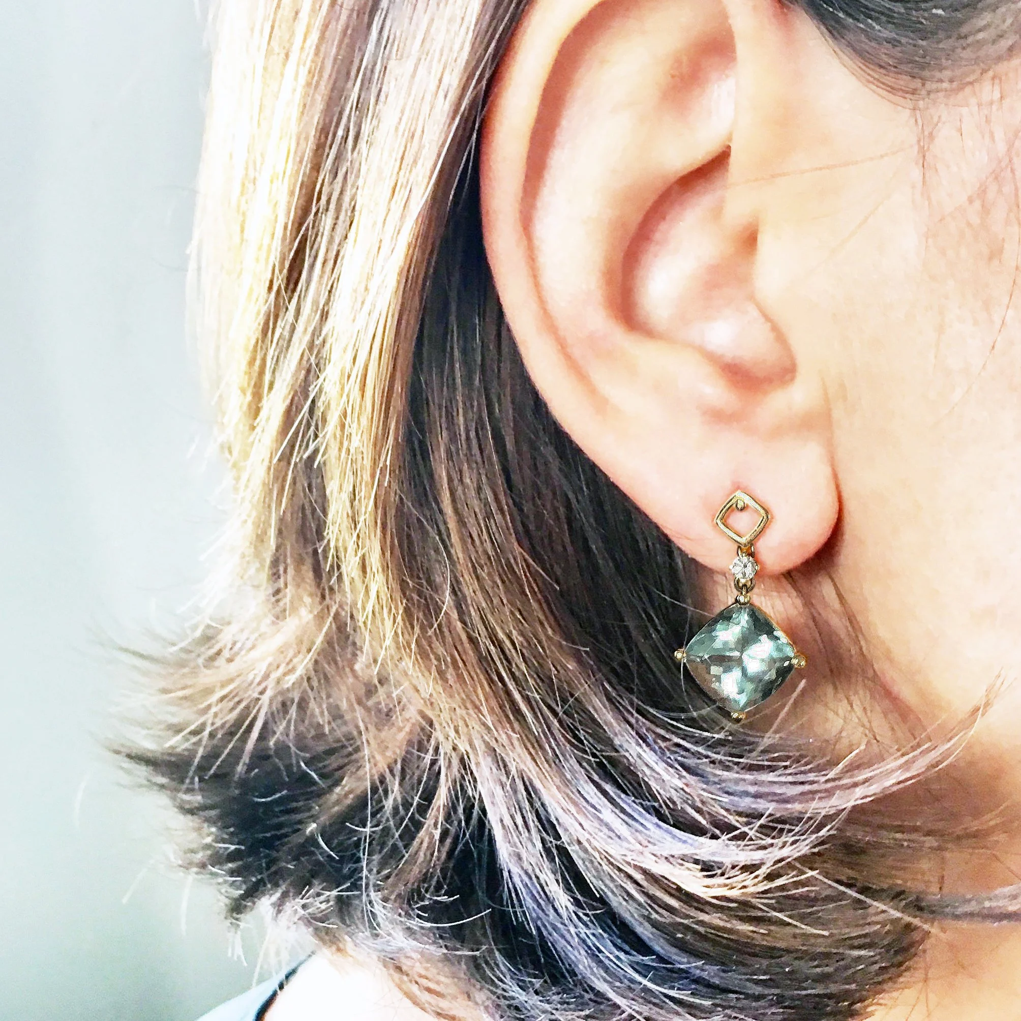 Close-up of a woman's ear wearing a dangling earring with a square gemstone and a small diamond above it, with her hair styled in light brown and gray layered hair.