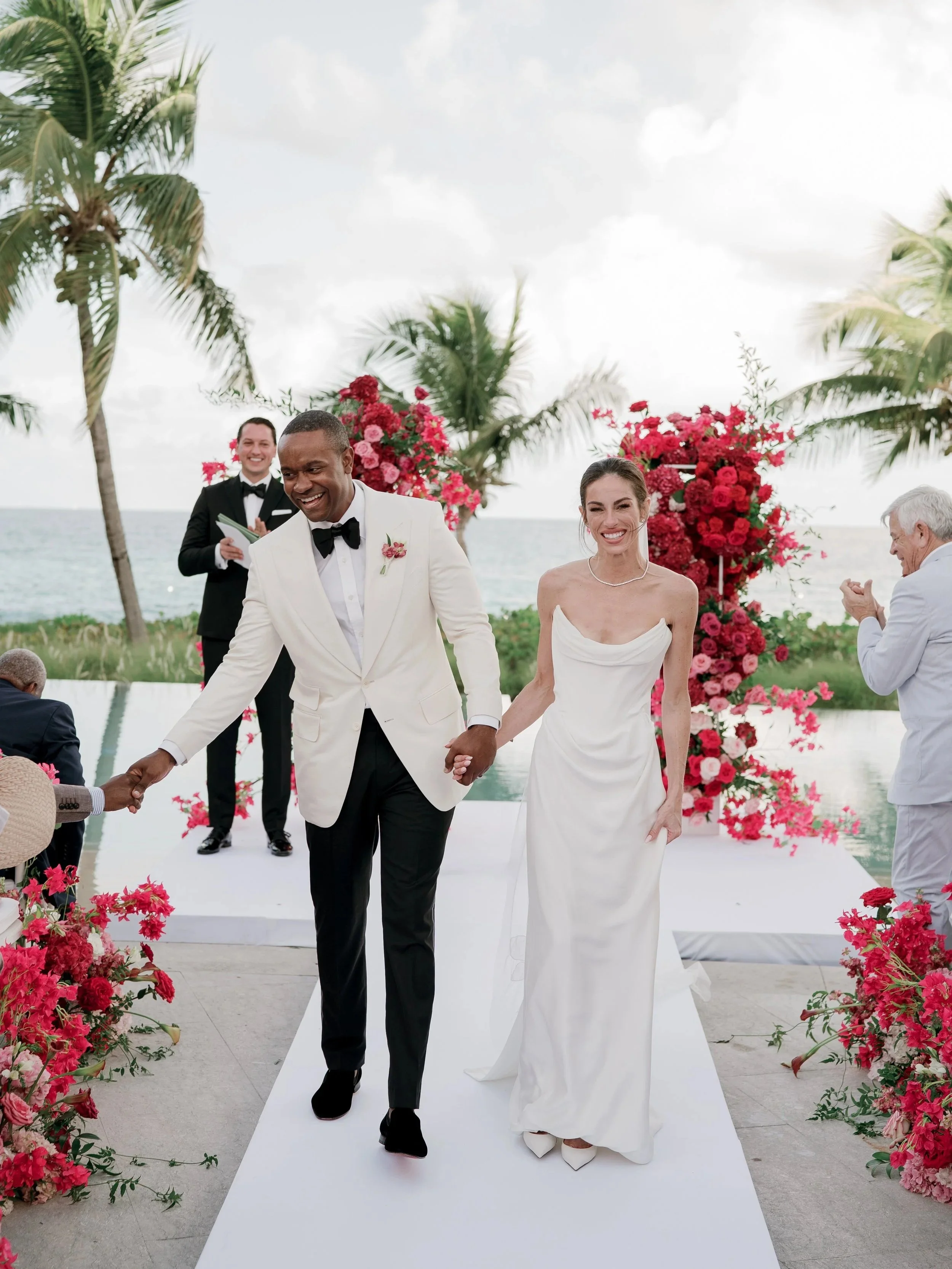 A wedding ceremony on a beach with the bride and groom holding hands and walking down an aisle decorated with pink and red flowers. The groom is wearing a white tuxedo with a black bow tie, and the bride is in a white wedding gown. A pastor or officiant and another man in a tuxedo are in the background, smiling.