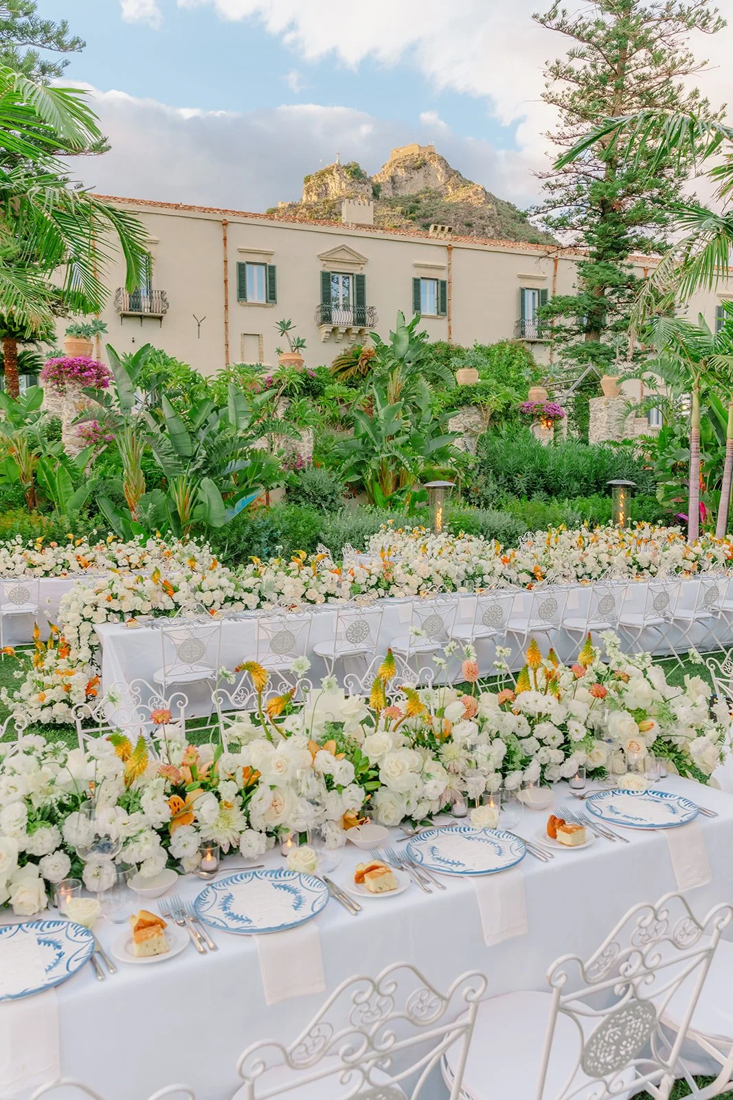 An outdoor wedding reception with long tables decorated with white and peach flowers, set with blue and white patterned plates, and surrounded by lush greenery and potted plants. A mountain with a castle on top is visible in the background.