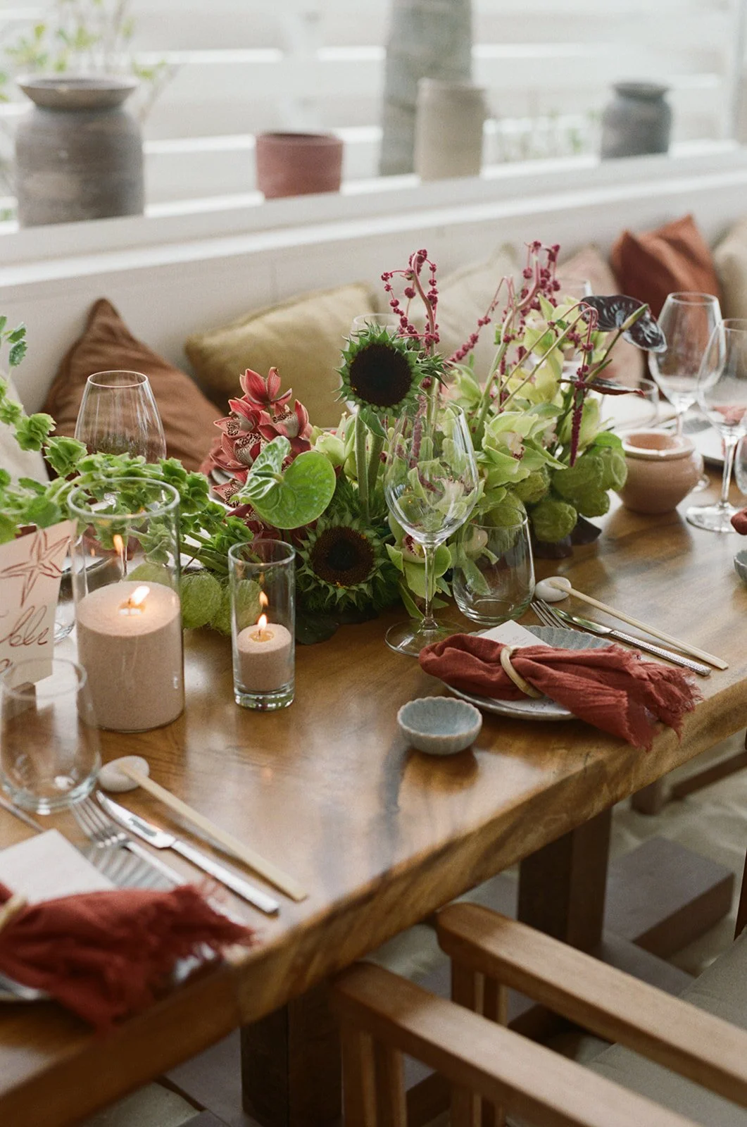 A decorated dining table with candles, wine glasses, floral centerpiece, and place settings with napkins and utensils in a bright, cozy room.