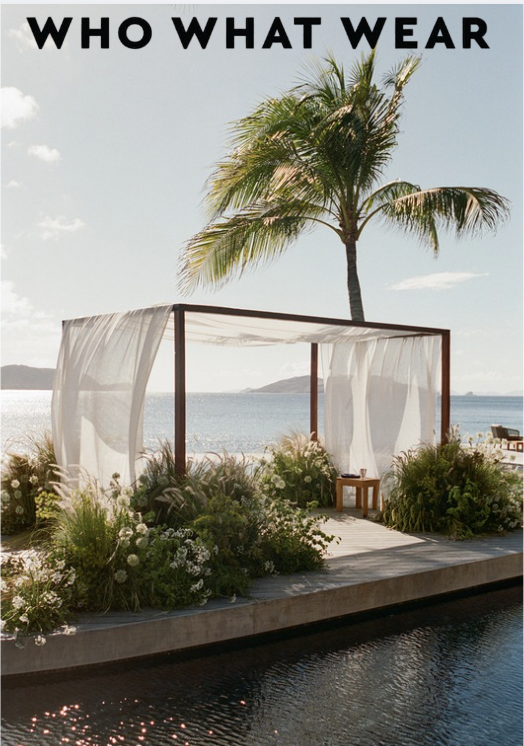 Beachside outdoor cabana with white curtains, surrounded by lush plants, in front of calm water and a palm tree under a clear sky. The text at the top reads "WHO WHAT WEAR."