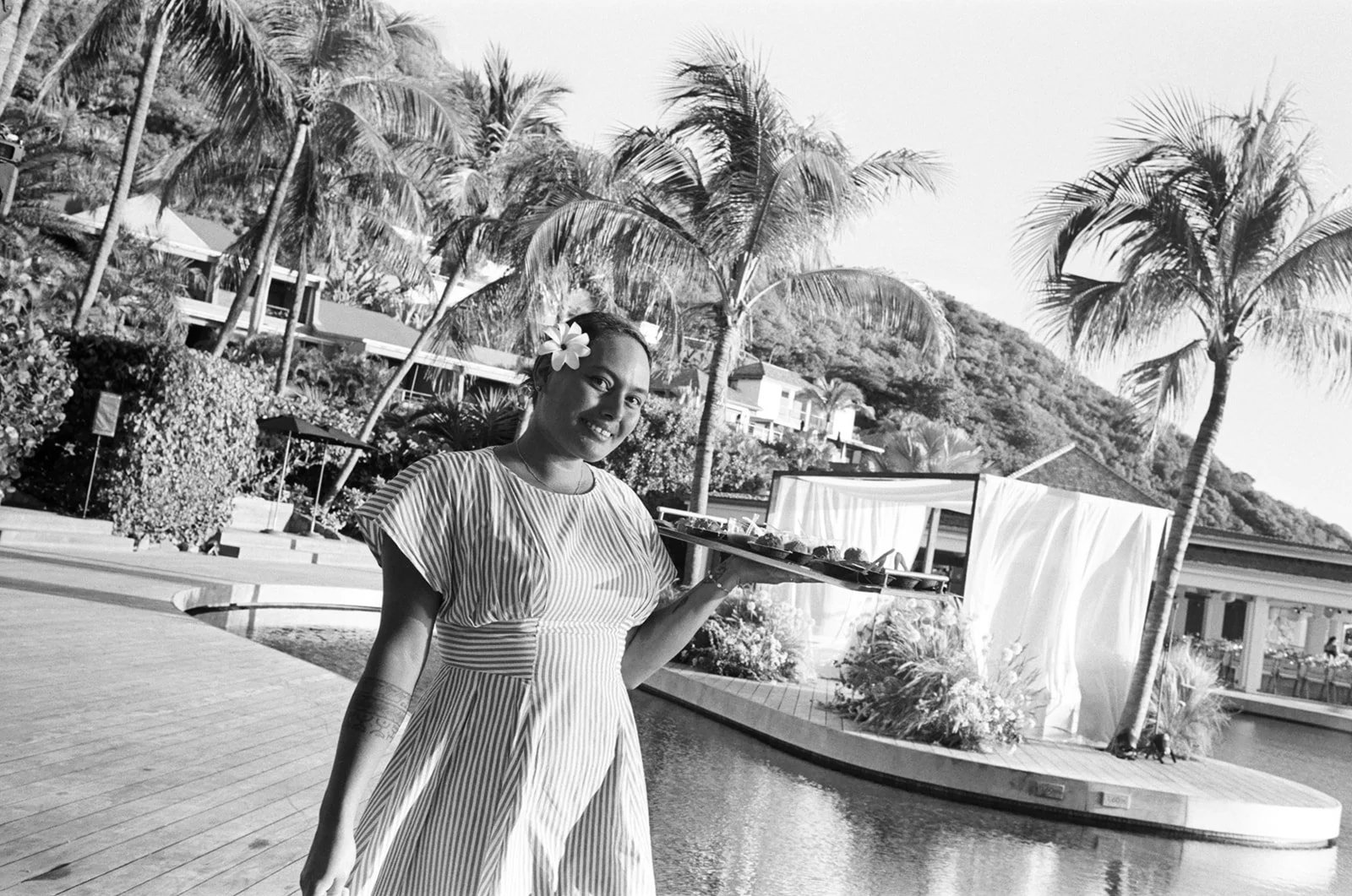 A woman wearing a striped dress with a flower in her hair, holding a tray of food outdoors with palm trees and a mountain in the background.