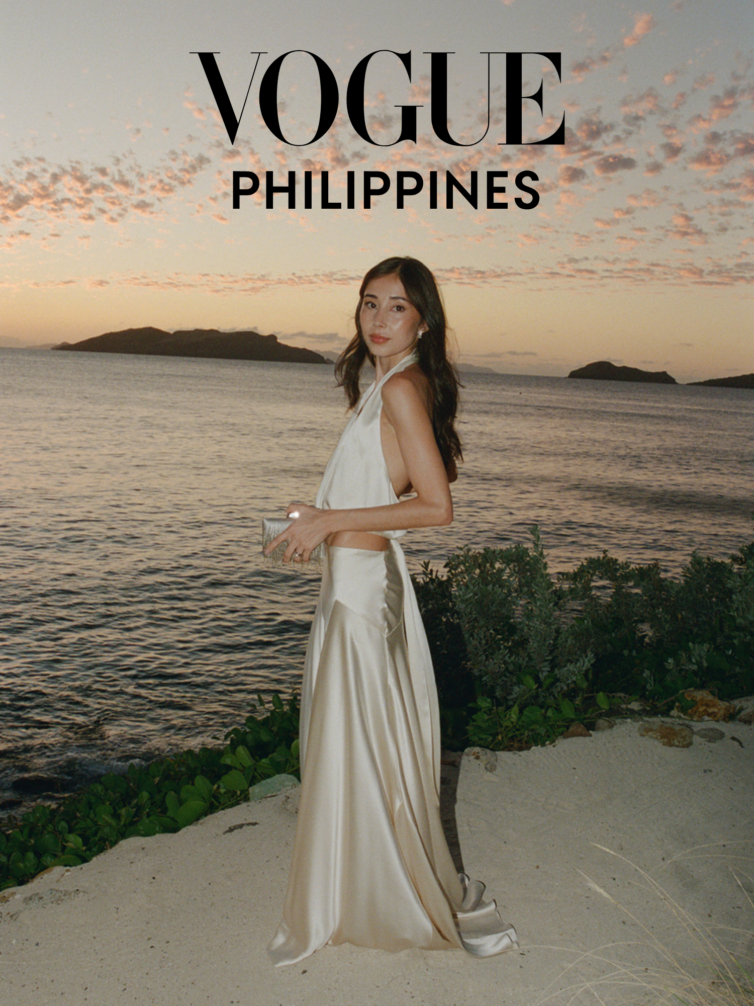 A woman in a white satin dress standing on the beach at sunset, with water and islands in the background.