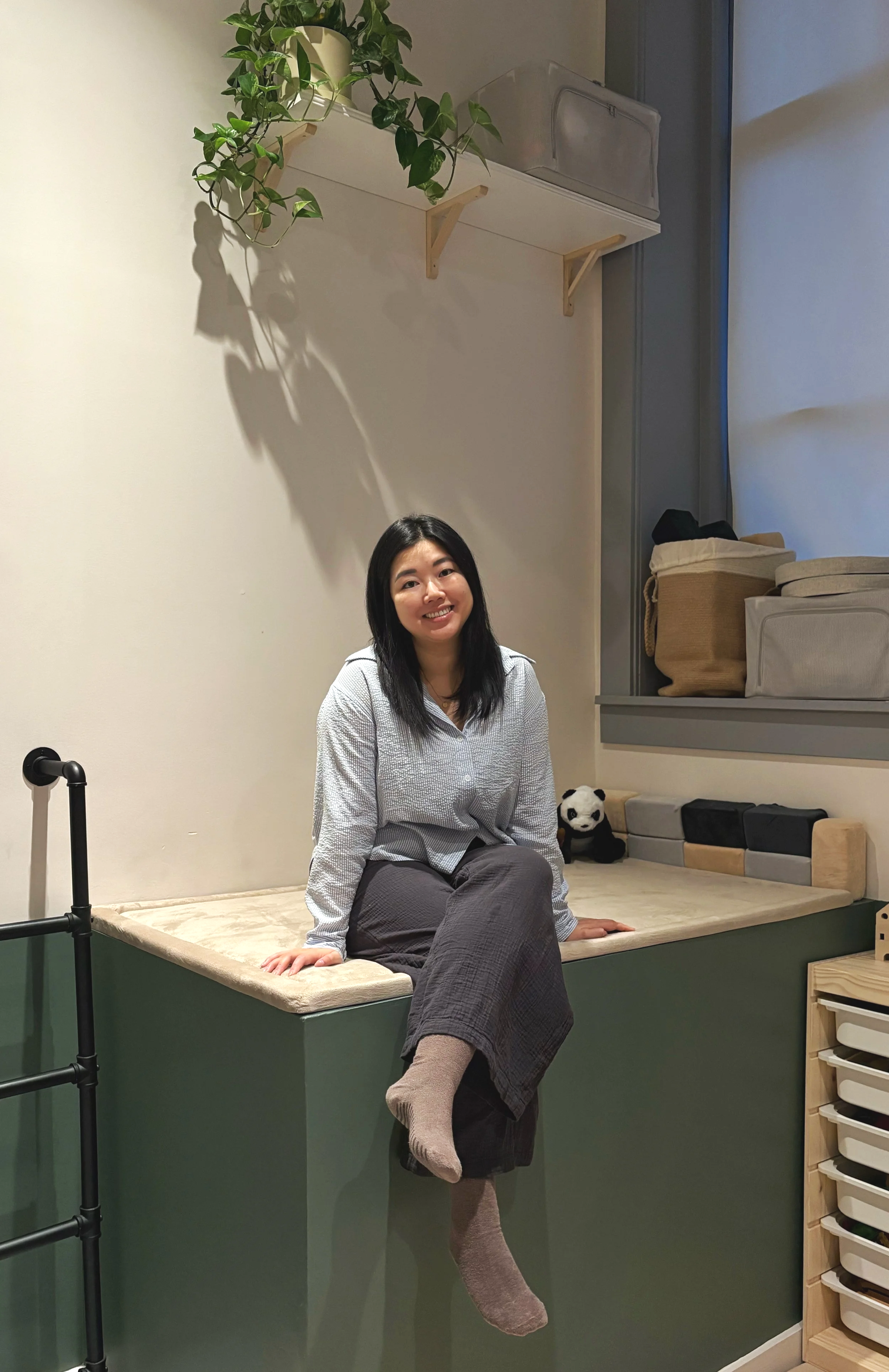 Diana, a speech-language pathologist at grow onwords, sitting on a built-in bench in a thoughtfully designed therapy space with sensory materials, storage shelves, and a hanging plant.