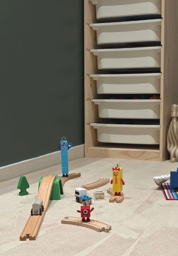 Wooden toy train tracks arranged on a light rug with small colorful number character figures standing on and around the tracks, set in a playroom corner with a wooden storage unit and green wall in the background.