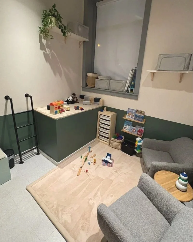 Cozy children’s playroom corner with green half-painted walls, a small window with books on the sill, wooden storage drawers, a low shelf with children’s books, and wooden train tracks and toys scattered across a light wood floor.