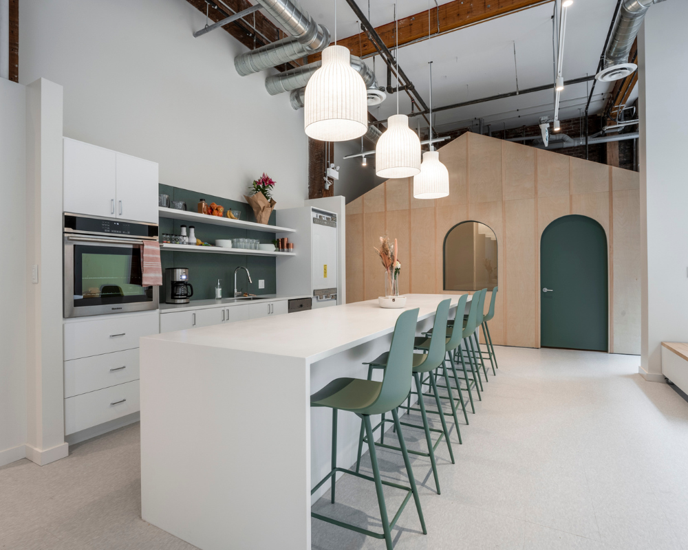 Modern common area at This World’s Ours Centre in Vancouver with a large kitchen island, green chairs, pendant lighting, and arched therapy room doorways.