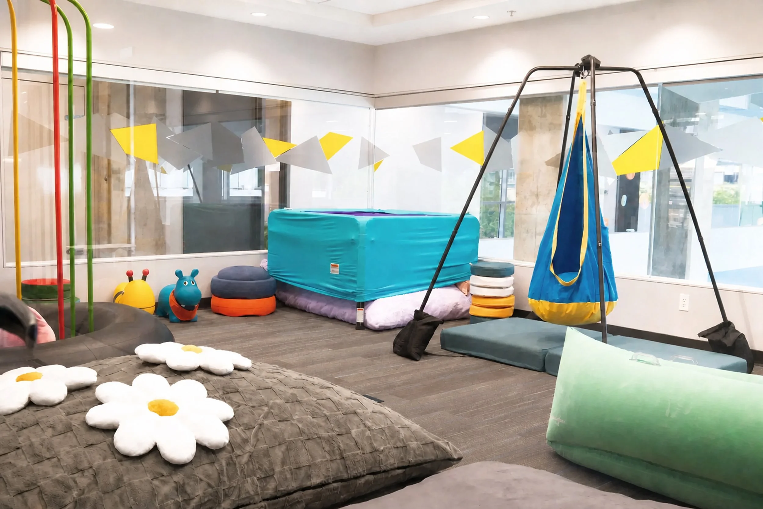 Sensory therapy room at The SPOT Pediatric Centrein Vancouver, BC, featuring a suspended platform swing, soft floor cushions, and floral and animal-shaped seating beneath large windows.