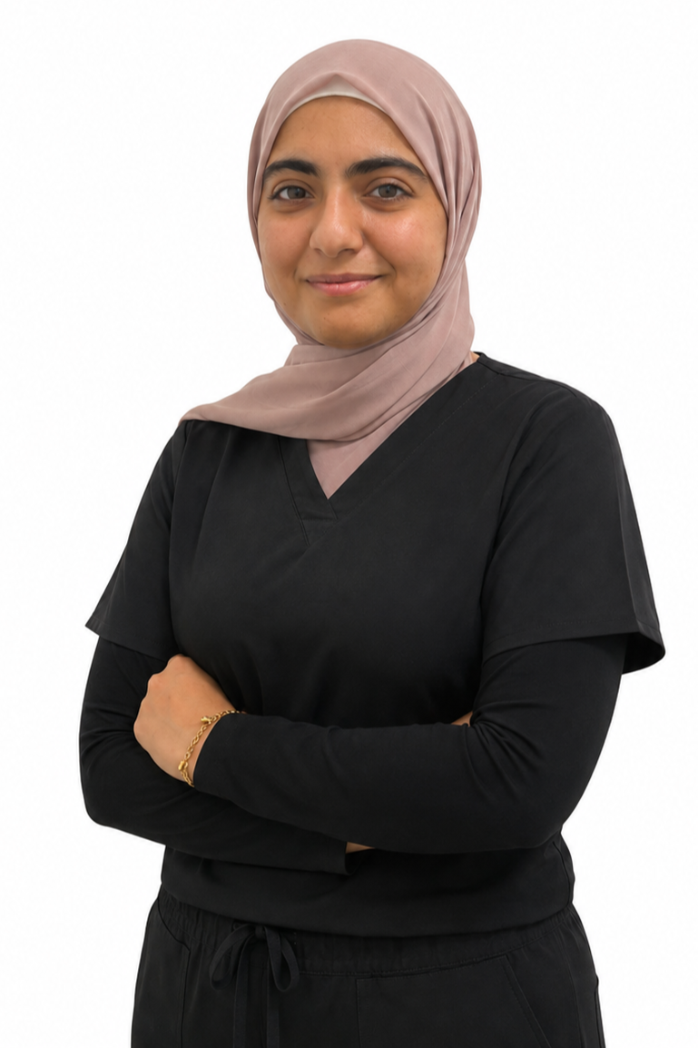 A woman wearing a light pink hijab and black medical scrubs, standing against a plain white background with arms crossed, smiling softly.