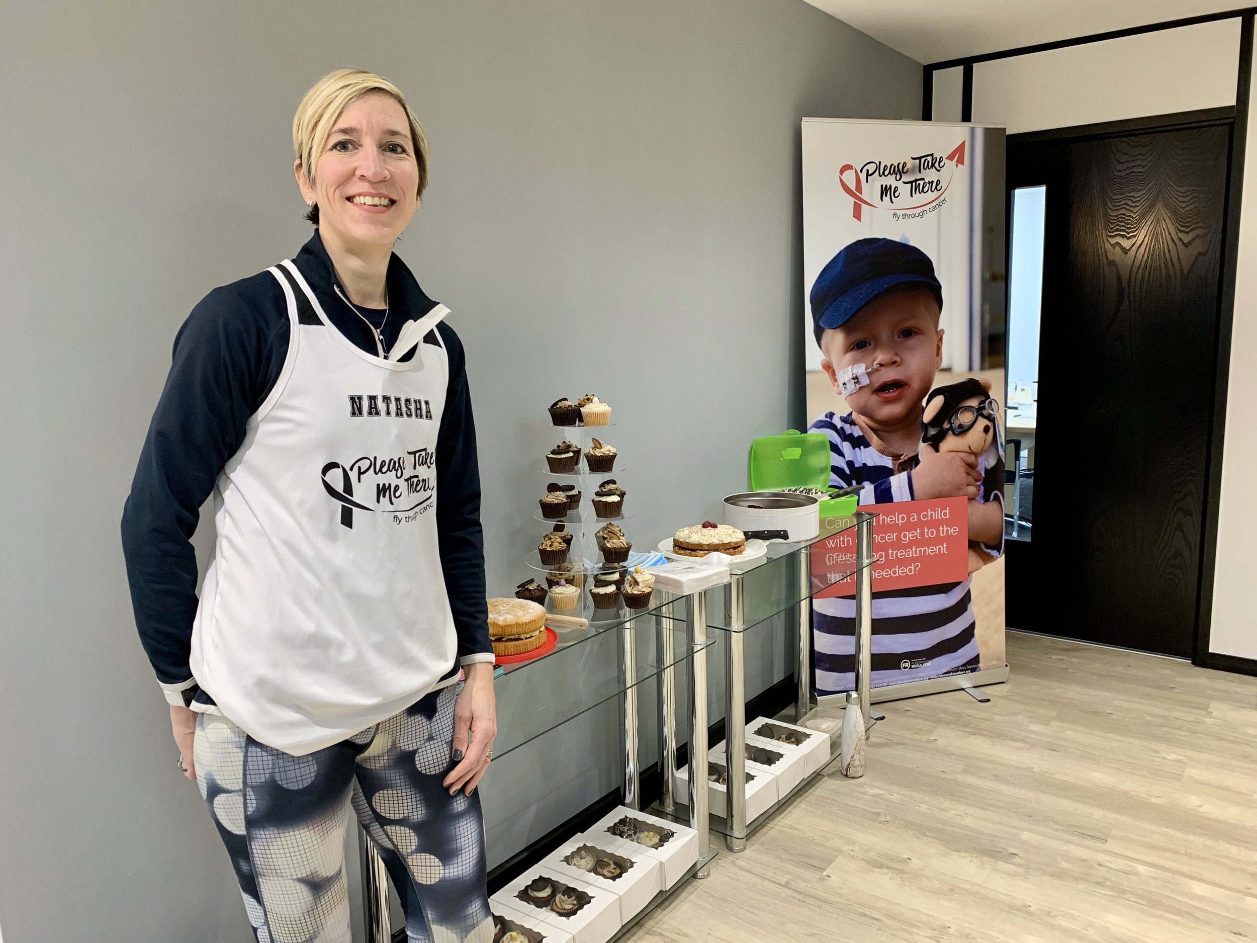 A woman wearing a white apron with the name Natasha and a black shirt underneath standing next to a table with cupcakes and a birthday cake. Behind her is a child in a striped shirt and blue hat holding a plush toy, with a cancer awareness banner in the background.